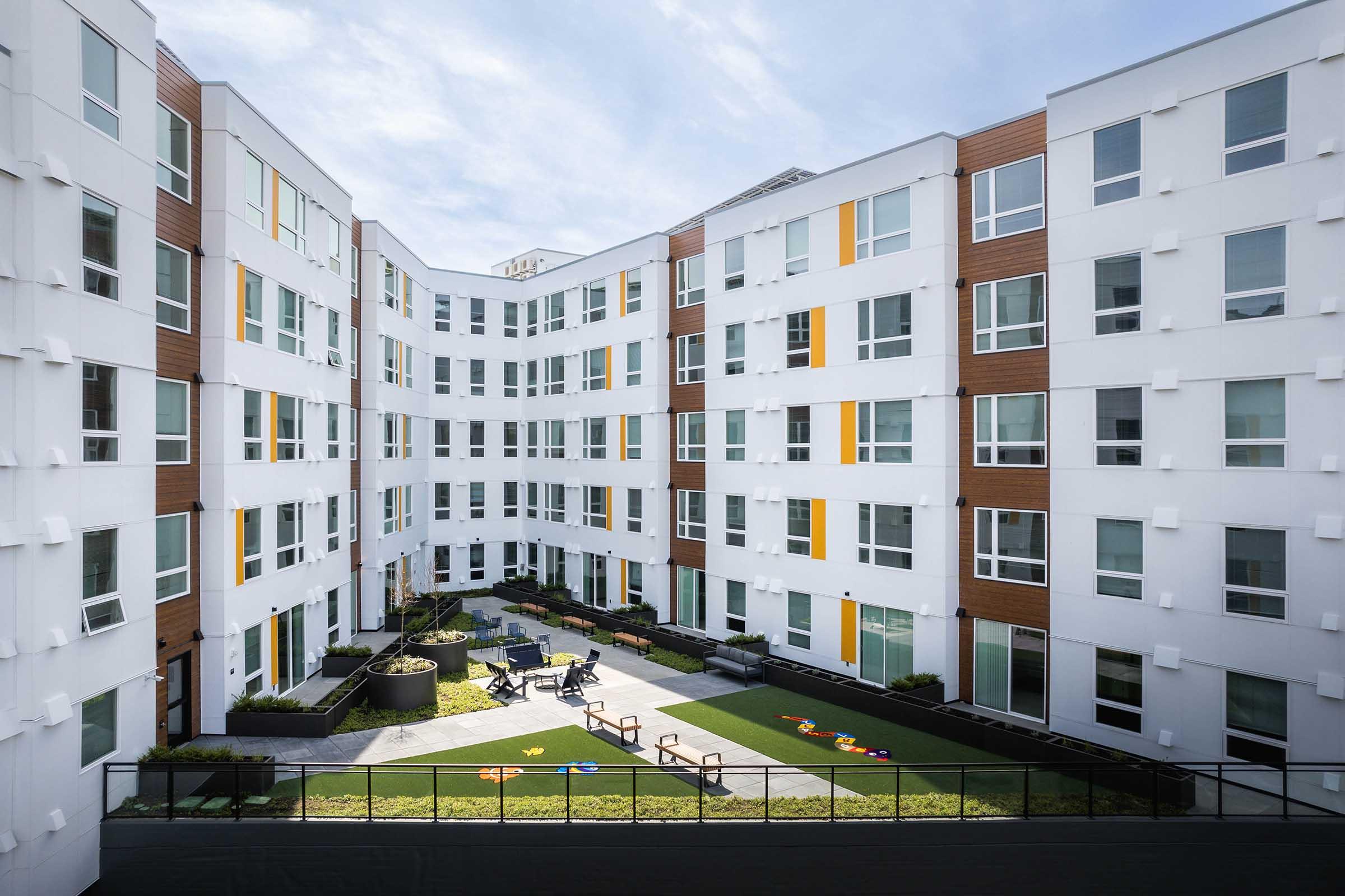 A modern apartment community with a central courtyard featuring green grass and seating areas. The building has a mix of white and wooden exterior elements, with large windows and colorful accents. The courtyard is designed for relaxation and outdoor activities, surrounded by the apartment units.