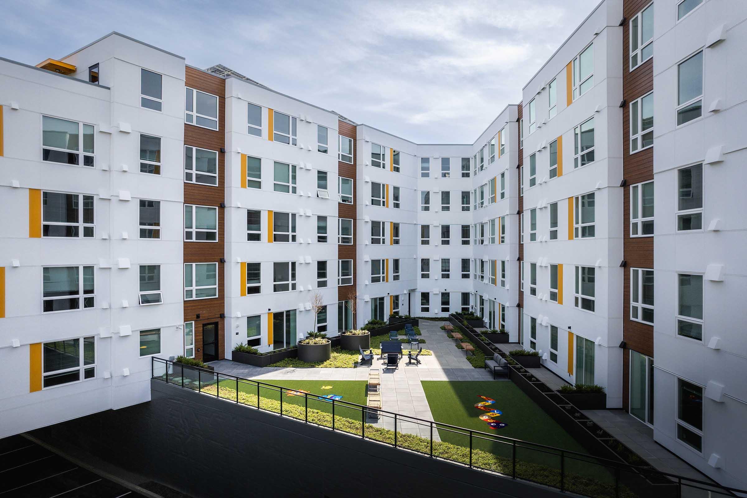 A modern apartment community featuring a central courtyard with green grass, seating areas, and colorful play equipment. The building has multiple stories with large windows and a mix of white and wooden facades, illuminated by natural light under a partly cloudy sky.