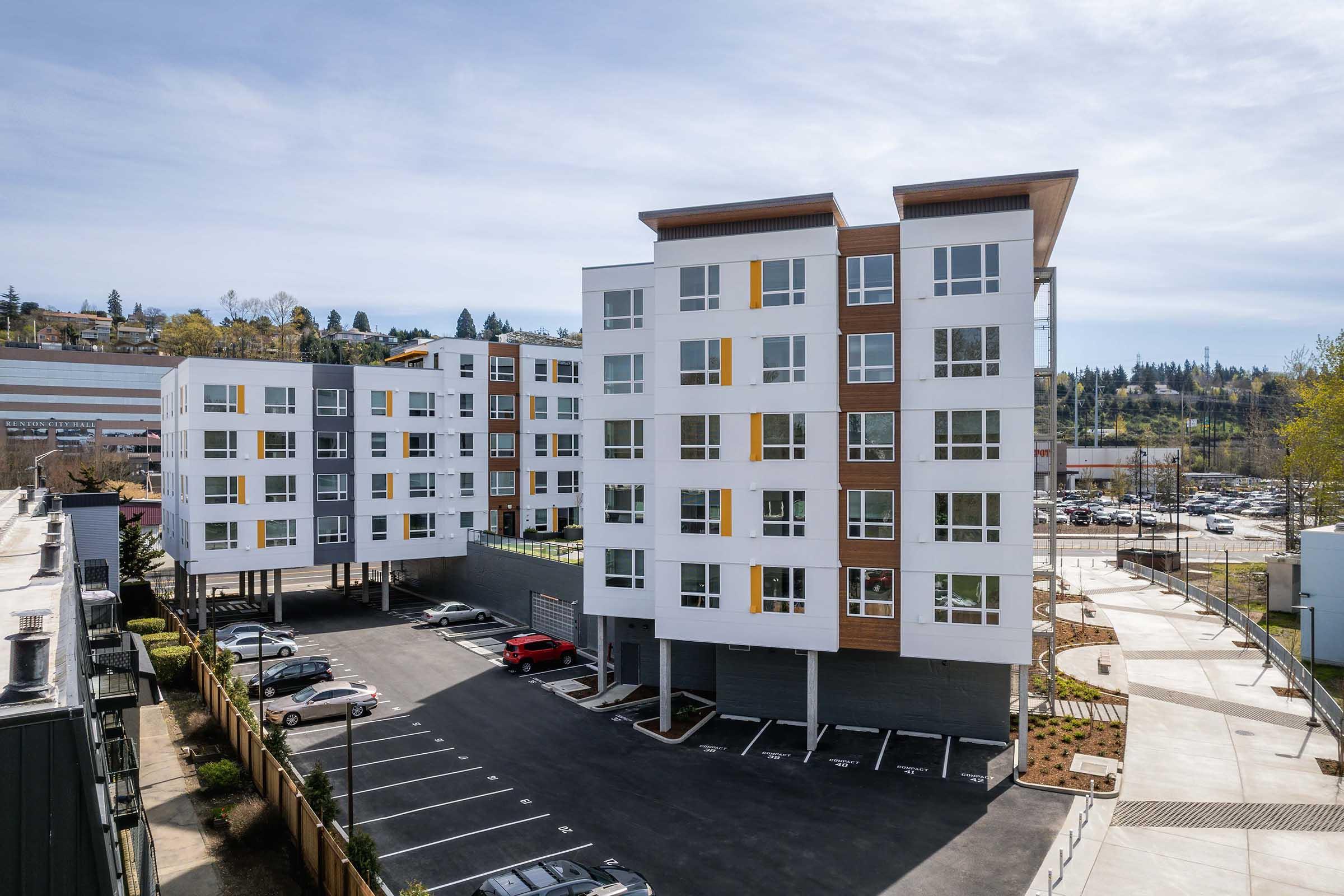 A modern residential building with a sleek design featuring multiple floors, large windows, and distinctive wooden accents. The structure is set in a parking lot with several parked cars, surrounded by landscaped areas. In the background, a cityscape and a parking facility are visible.