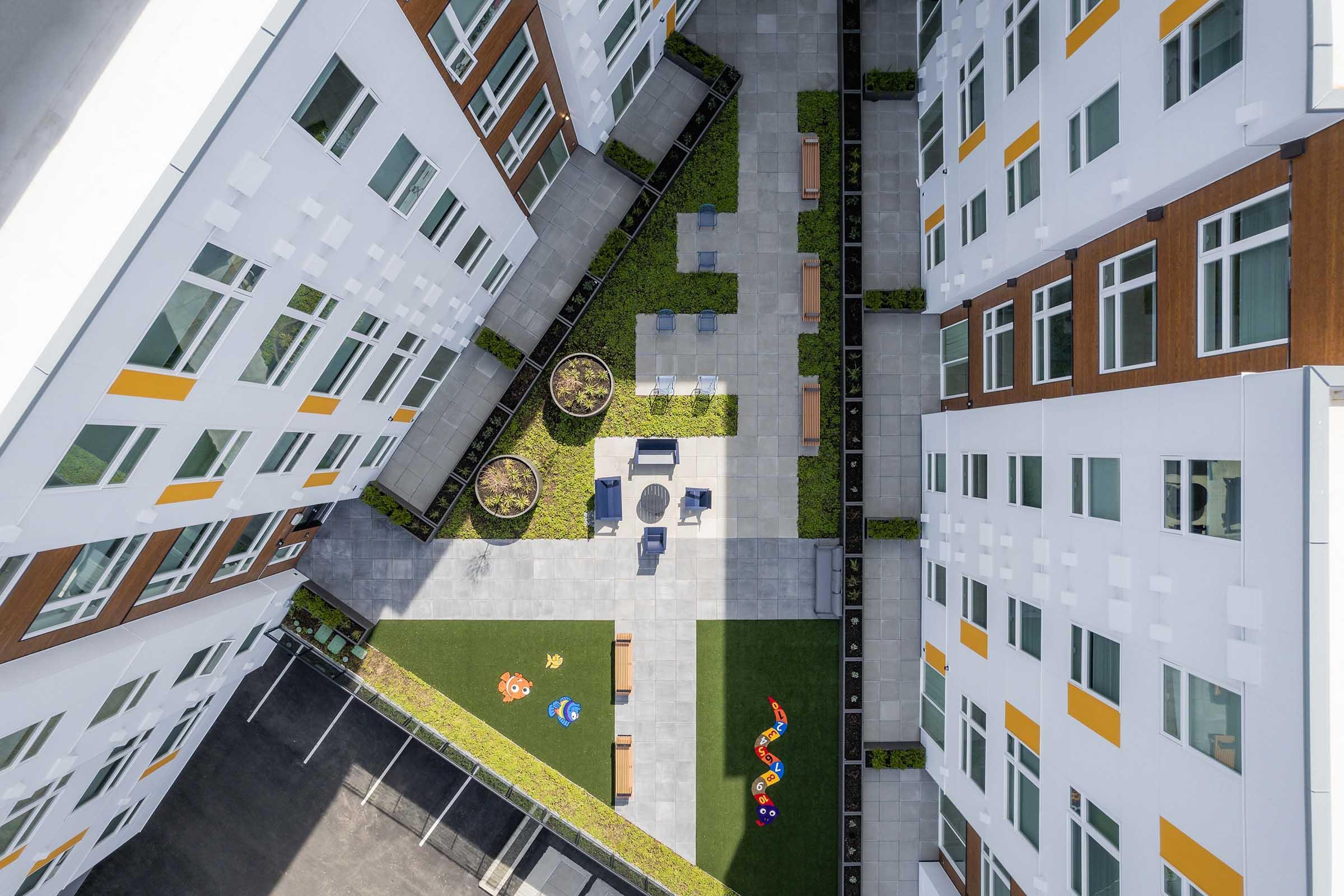 Aerial view of a modern building complex featuring a landscaped courtyard with seating areas, grassy patches, and children's toys scattered on the ground. The surrounding buildings are white with wooden accents, creating a vibrant and welcoming environment.