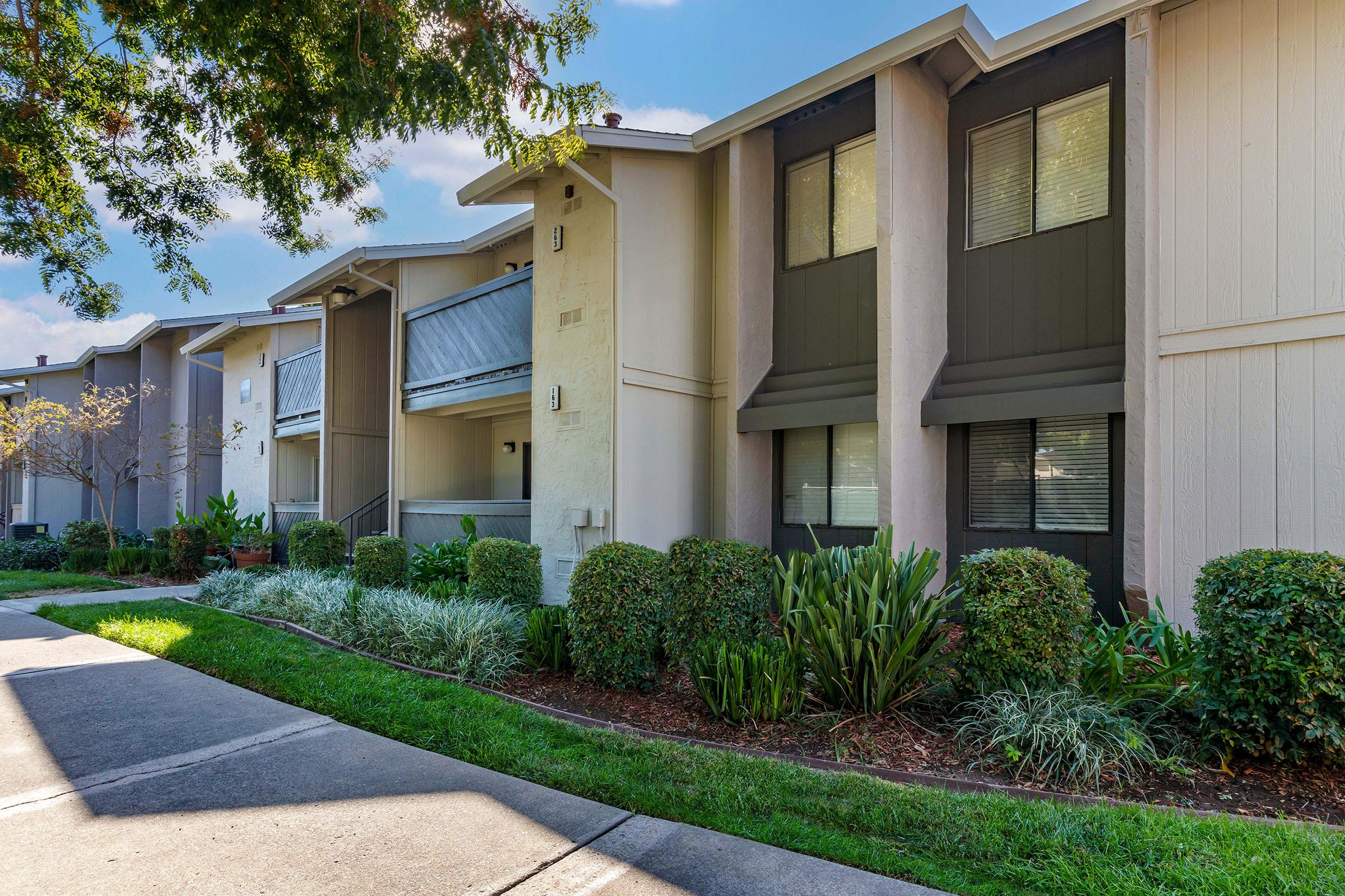 A view of a modern apartment complex featuring multiple two-story buildings with balconies, well-manicured landscaping including shrubs and grass, and a paved walkway. The scene is bright and inviting, with a clear blue sky in the background.