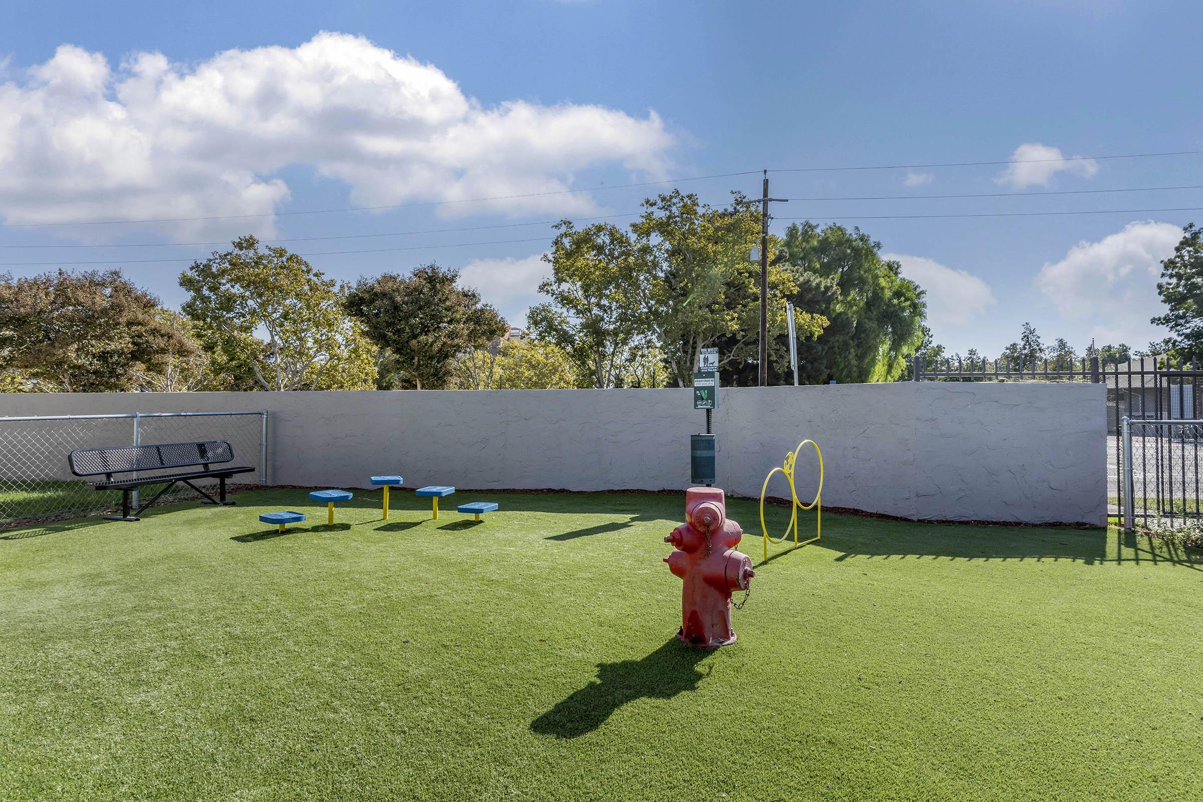 A vibrant outdoor dog park featuring green artificial turf, a red fire hydrant, yellow agility hoops, and blue benches. The area is enclosed with a white wall and a chain-link fence, surrounded by trees and under a bright blue sky with fluffy clouds.