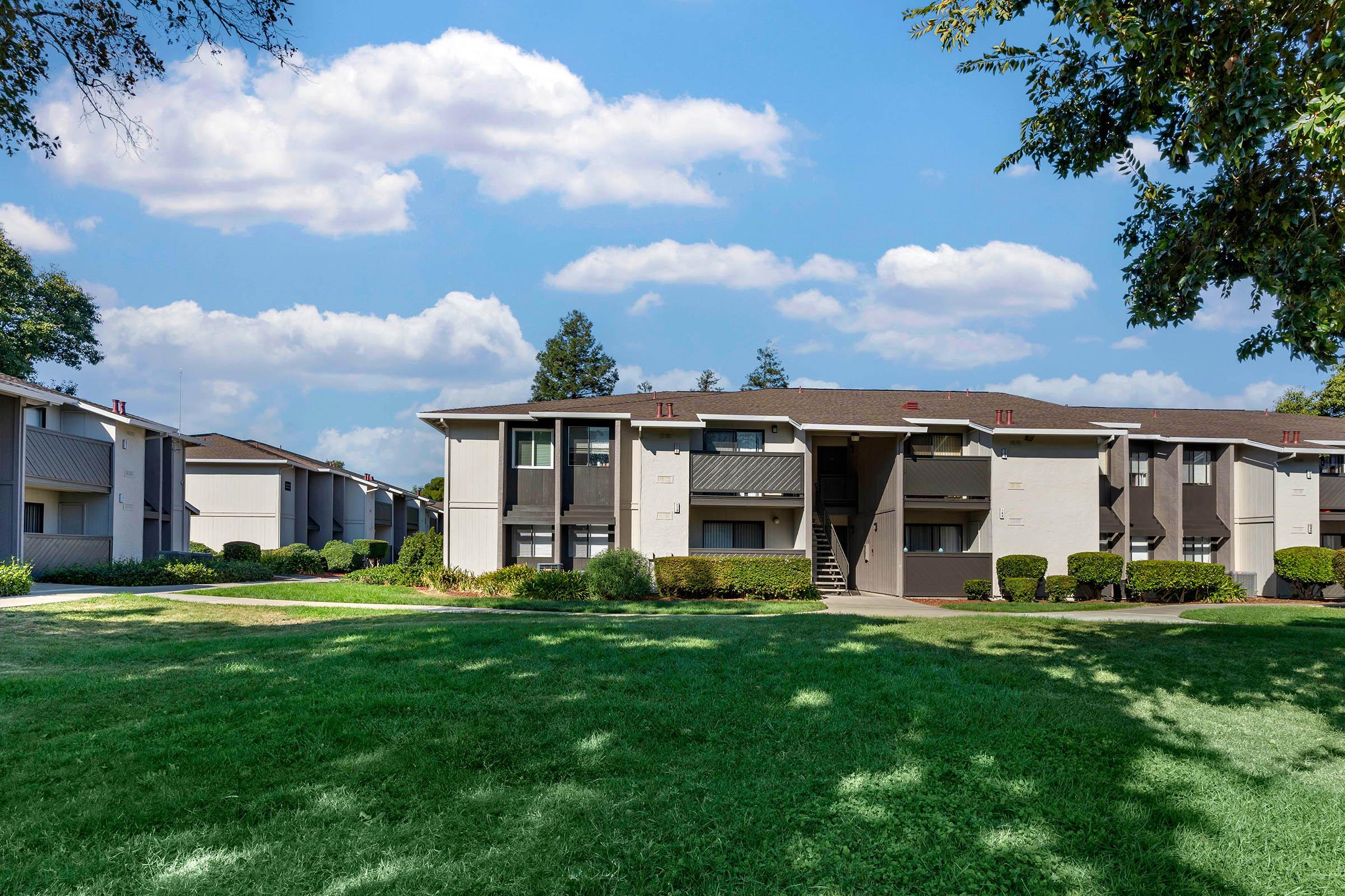 A brightly lit view of an apartment complex with two-story buildings surrounded by green lawns and trees under a partly cloudy blue sky. The well-maintained landscaping features shrubs and a pathway leading to the entrance of the buildings.