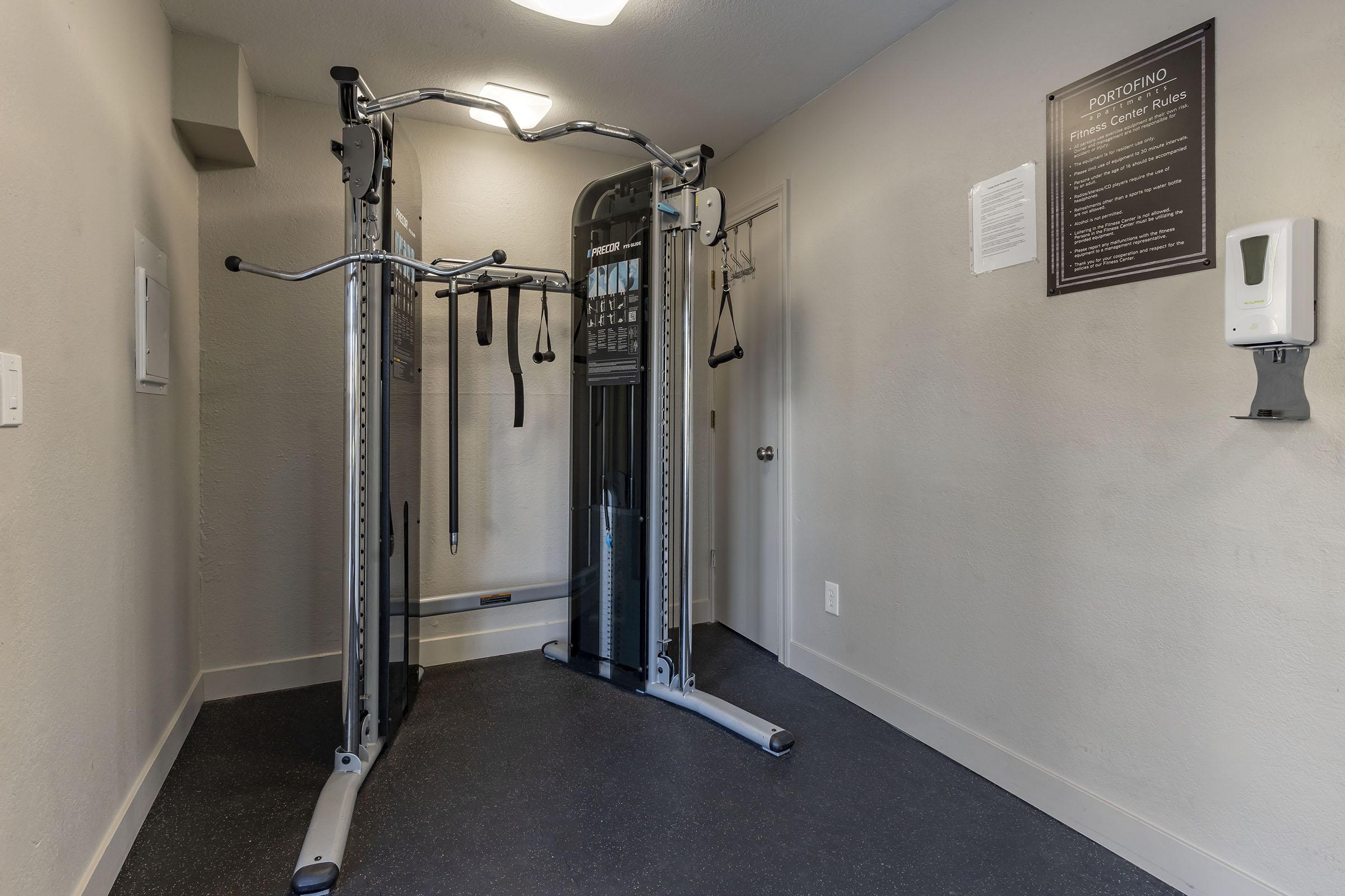 A small exercise room featuring a multi-gym machine with adjustable cables and resistance bands. There is a wall-mounted hand sanitizer dispenser and a notice on the wall with fitness information. The room is well-lit, with gray walls and a dark flooring surface.