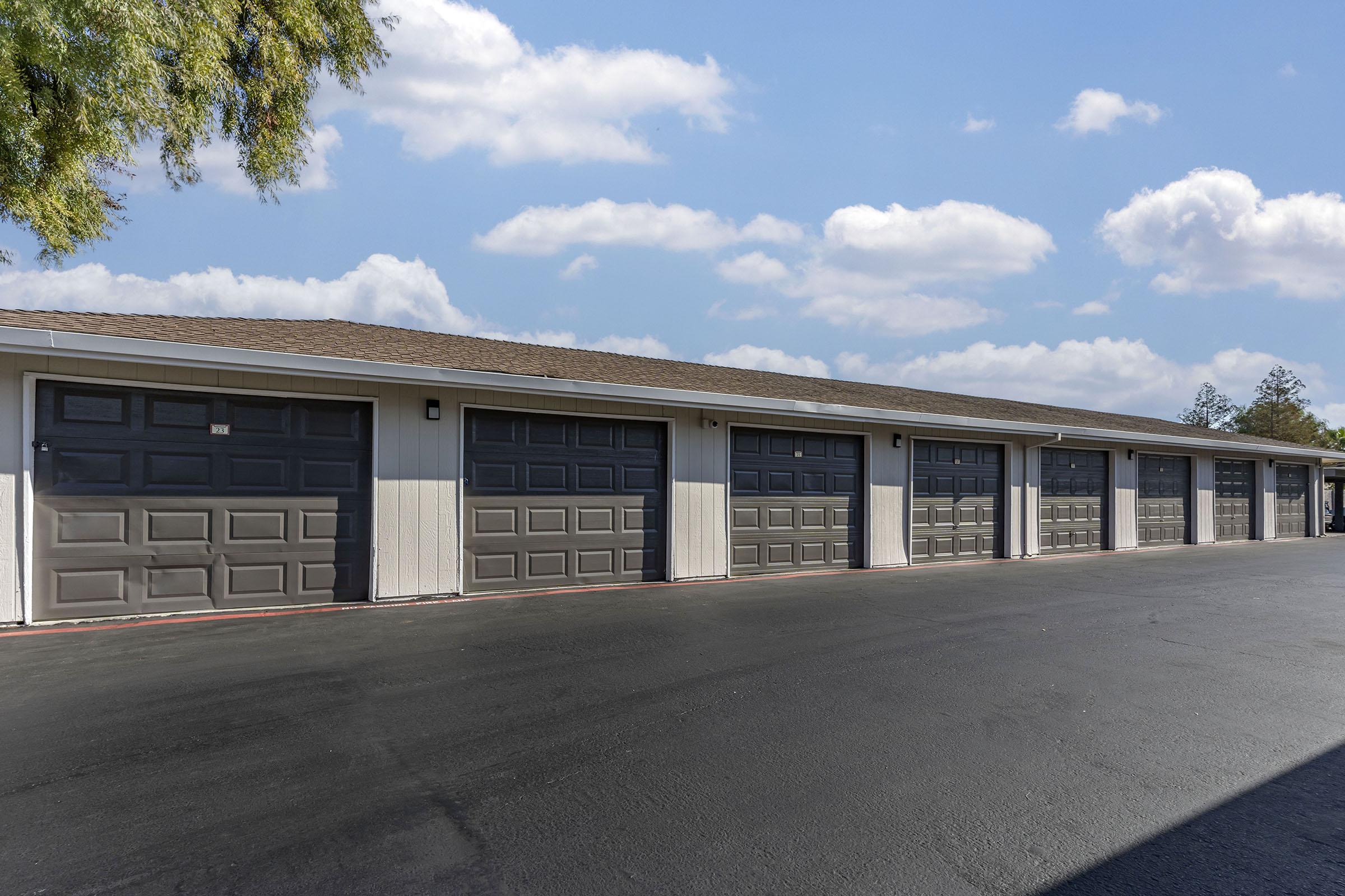 A row of several closed gray garage doors in a well-maintained outdoor area. The building has a light-colored exterior with a sloped roof, and the sky above is partly cloudy with a few scattered clouds. The asphalt driveway in front is clean and newly paved.