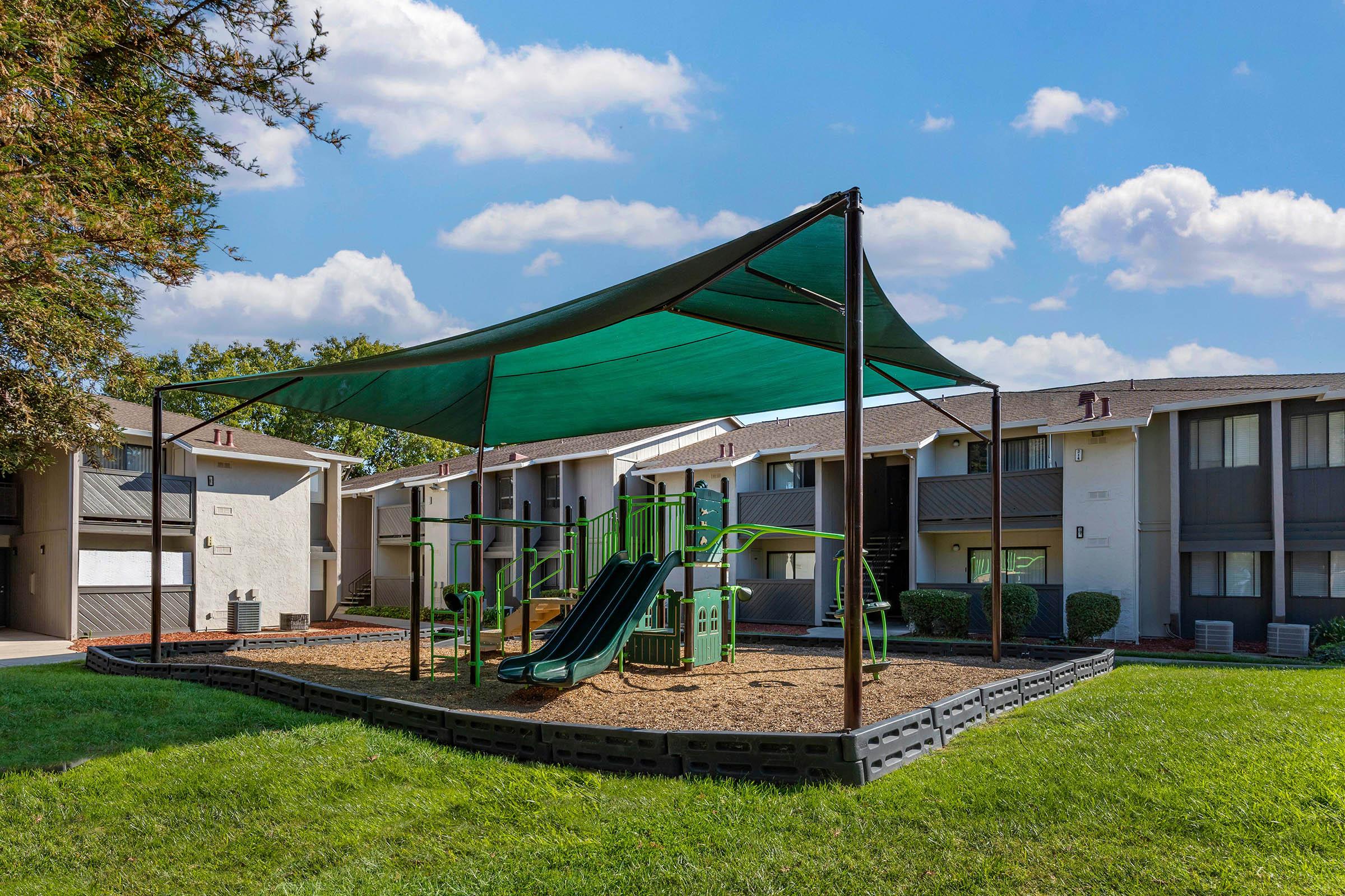 A playground featuring green equipment, including slides and climbing structures, is shaded by a large canopy. It is situated in a grassy area surrounded by residential apartment buildings under a partly cloudy sky.
