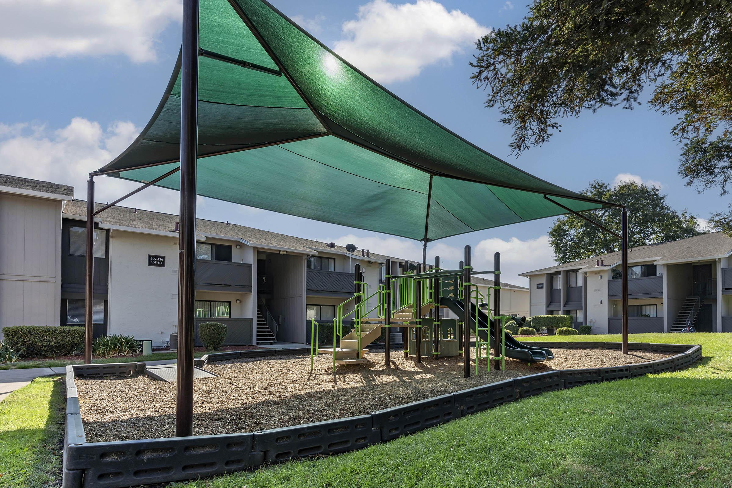 Playground area with a shaded structure, featuring a slide and climbing equipment, surrounded by grassy areas and apartment buildings in the background. The scene is set under a blue sky with scattered clouds.