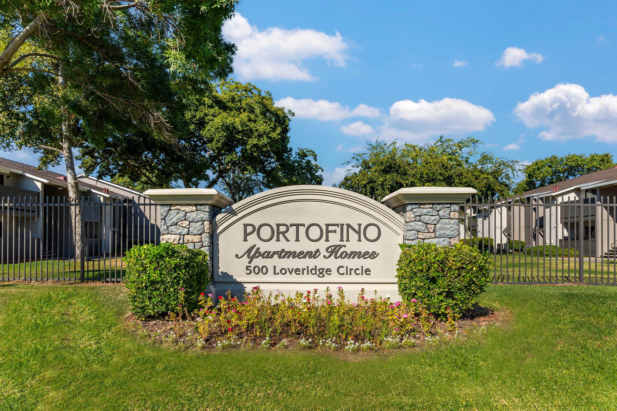 Sign for Portofino Apartment Homes, displaying the name and address "500 Loveridge Circle." The sign features a stone base and is surrounded by manicured grass and landscaping, with trees and a clear blue sky in the background.