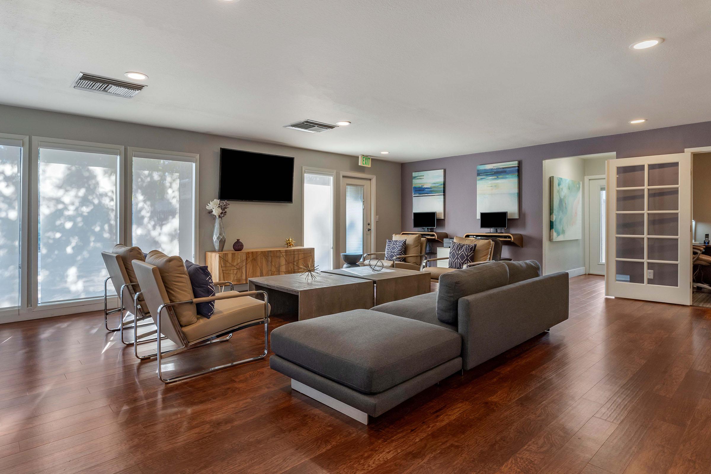 A modern living room featuring hardwood flooring, a sectional sofa, a coffee table, and sleek chairs. The walls are painted in soft tones, and there are large windows allowing natural light. A flat-screen TV is mounted on one wall, and art pieces are displayed. Two workstations are visible in a nearby room.