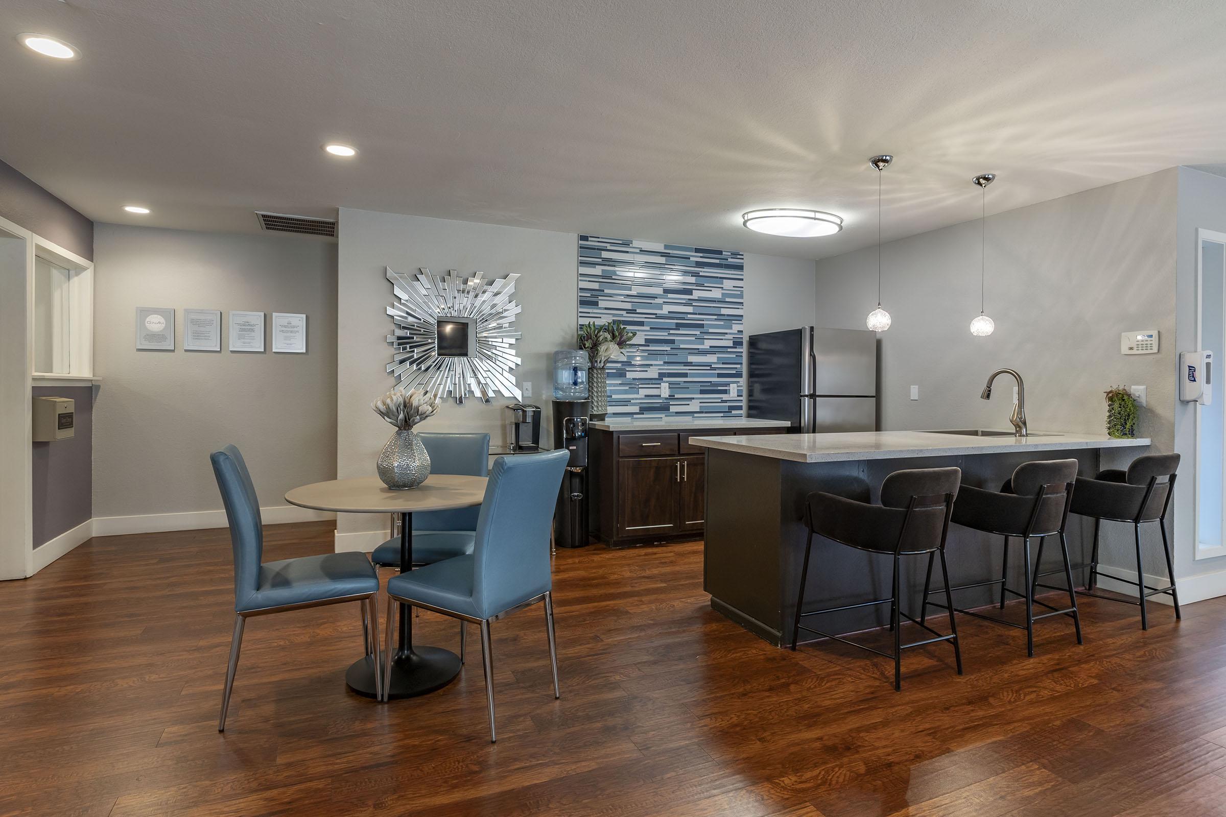 Modern kitchen and dining area featuring a round table with two blue chairs, a dark countertop island with bar stools, and a decorative wall mirror. The backsplash displays blue tiles, with appliances and cabinets visible in the background. The flooring is dark wood, creating a warm ambiance.
