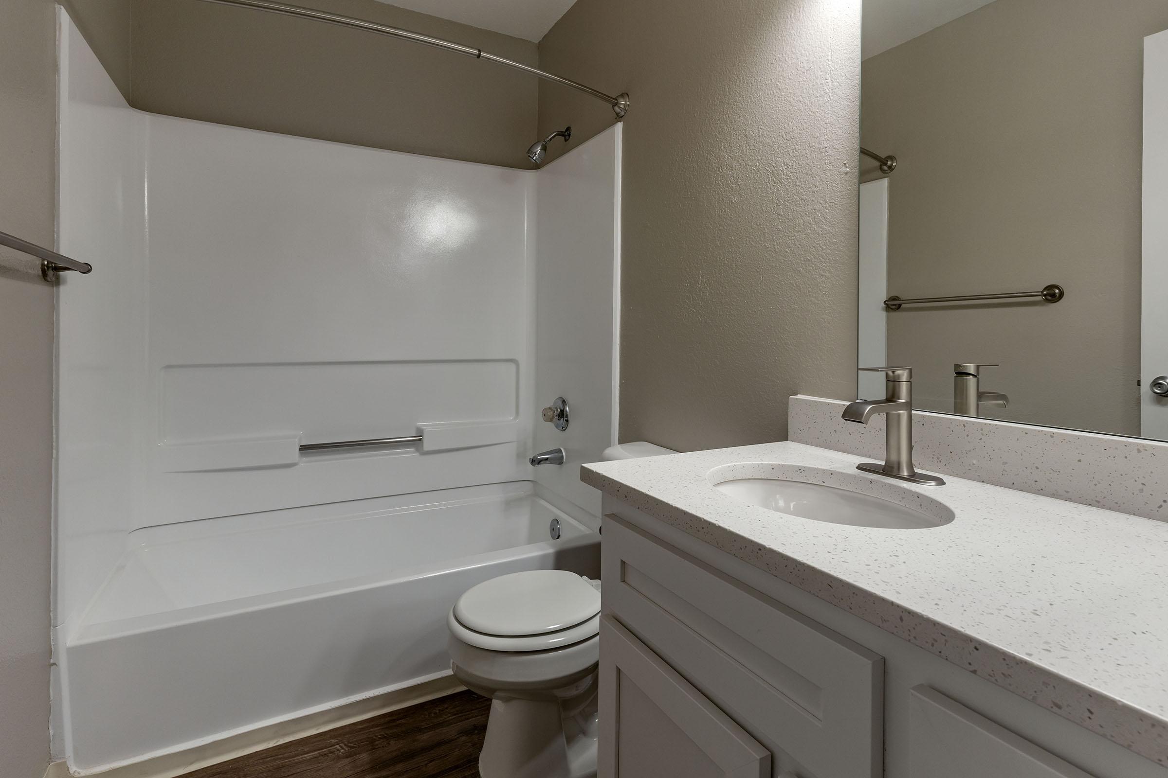 A modern bathroom featuring a white tub and shower combination, a light-colored countertop with a sink, and a mirror. The walls are painted in a muted tone, and there are chrome fixtures and a towel rack. The floor appears to be wood-like, adding warmth to the space.