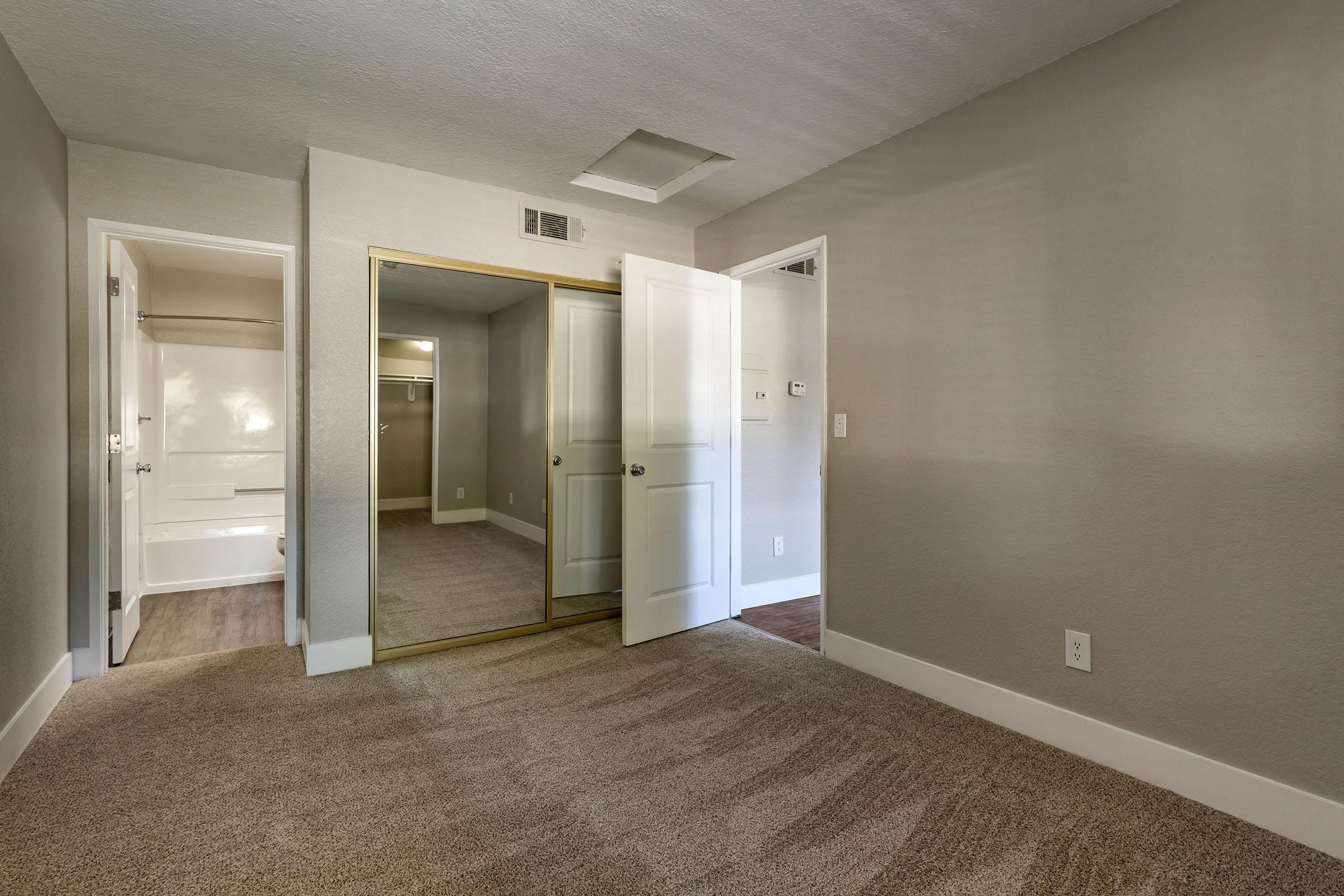 A well-lit, neutral-colored room with light gray walls and carpeted flooring. There are two open doors: one leading to a closet with mirrored sliding doors, and another leading to a bathroom or additional space. The room appears spacious and clean, suitable for various uses.
