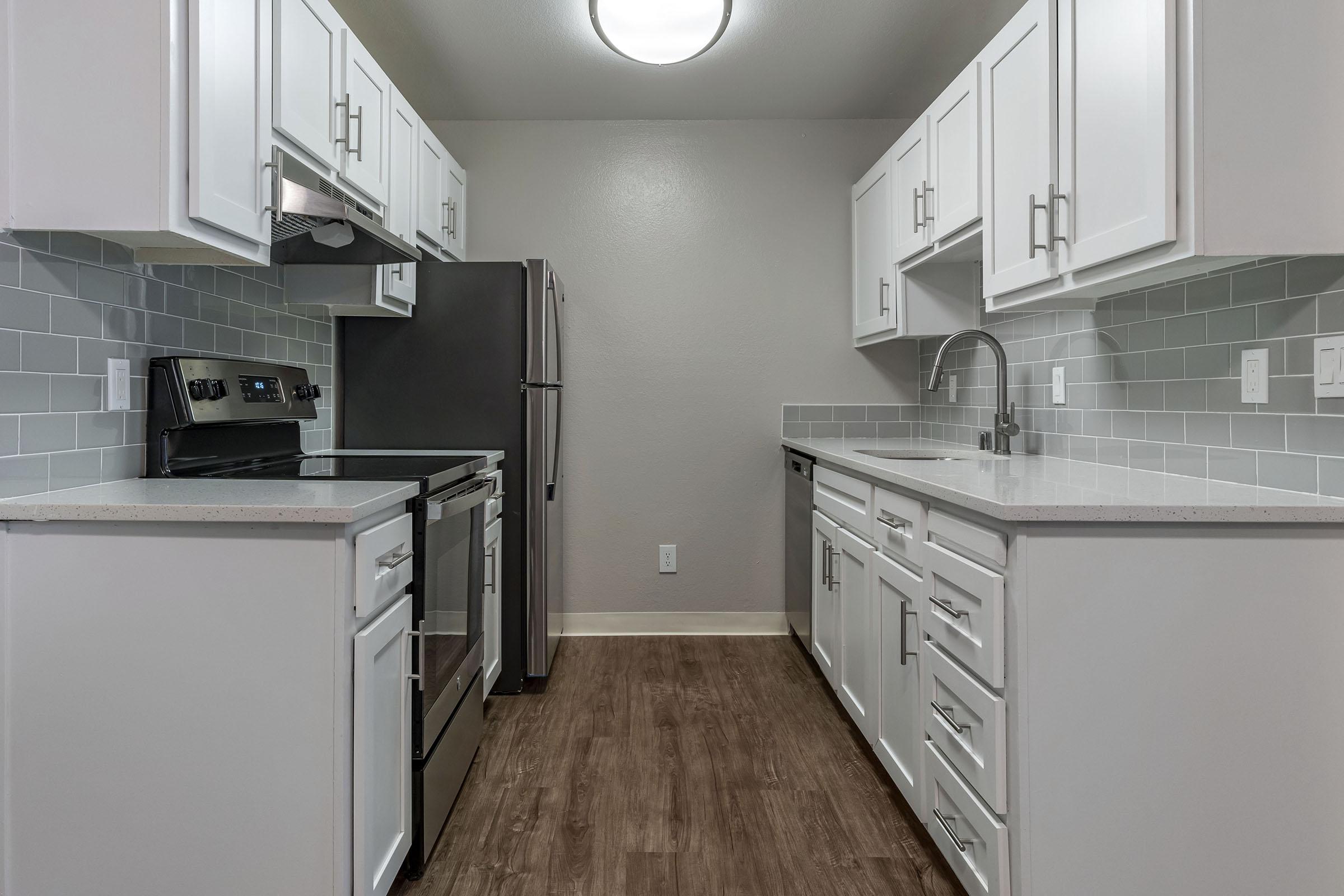 Modern kitchen featuring white cabinets, stainless steel appliances, a gray backsplash, and wood-style flooring. The layout includes a stove, oven, refrigerator, and sink with a faucet. Bright overhead lighting enhances the space, creating a clean and inviting atmosphere.