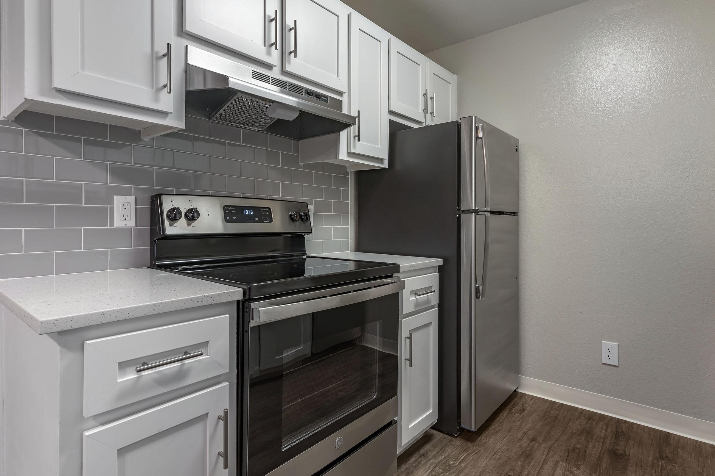 Modern kitchen with white cabinetry, stainless steel appliances including an oven and refrigerator, and a light gray tiled backsplash. The countertop is a solid surface. The flooring is dark wood, creating a contemporary and clean aesthetic.