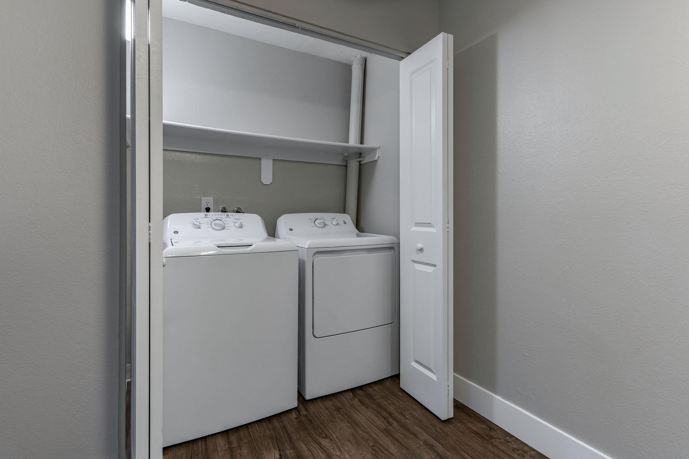 A small laundry room featuring a stacked washer and dryer located in a closet. The walls are painted in a light color, and there is a shelf above the appliances. The floor is made of wooden planks, and a white door leads to the room.