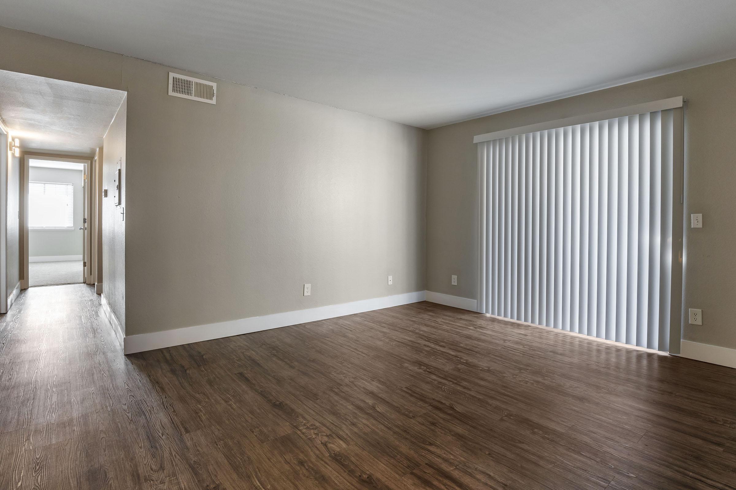 A spacious, empty living room with laminate wood flooring and light gray walls. On the right, there's a large window covered with vertical blinds that allow natural light to enter. A hallway is visible on the left, leading to another area of the space, enhancing the open and inviting atmosphere.