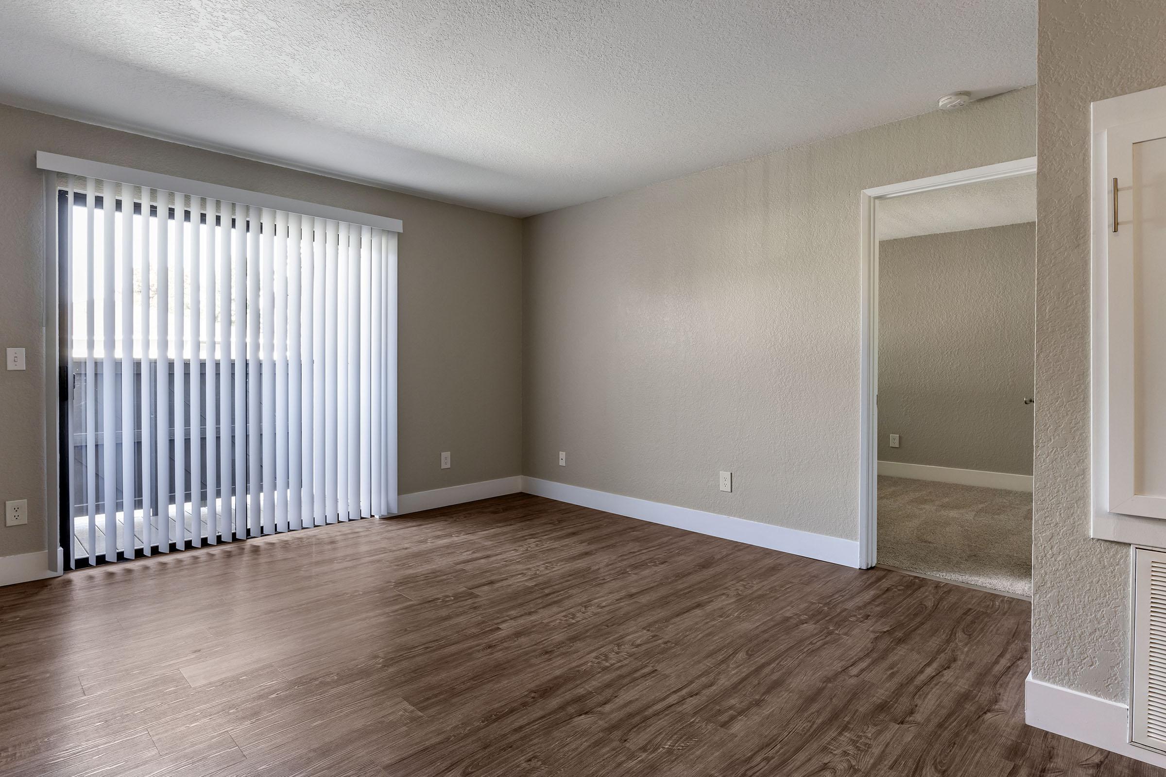 A spacious and empty room with light brown wood-like flooring, painted light gray walls, and vertical blinds on a large window. There is an open doorway leading to another area, with no furniture present, creating a clean and minimalist look.