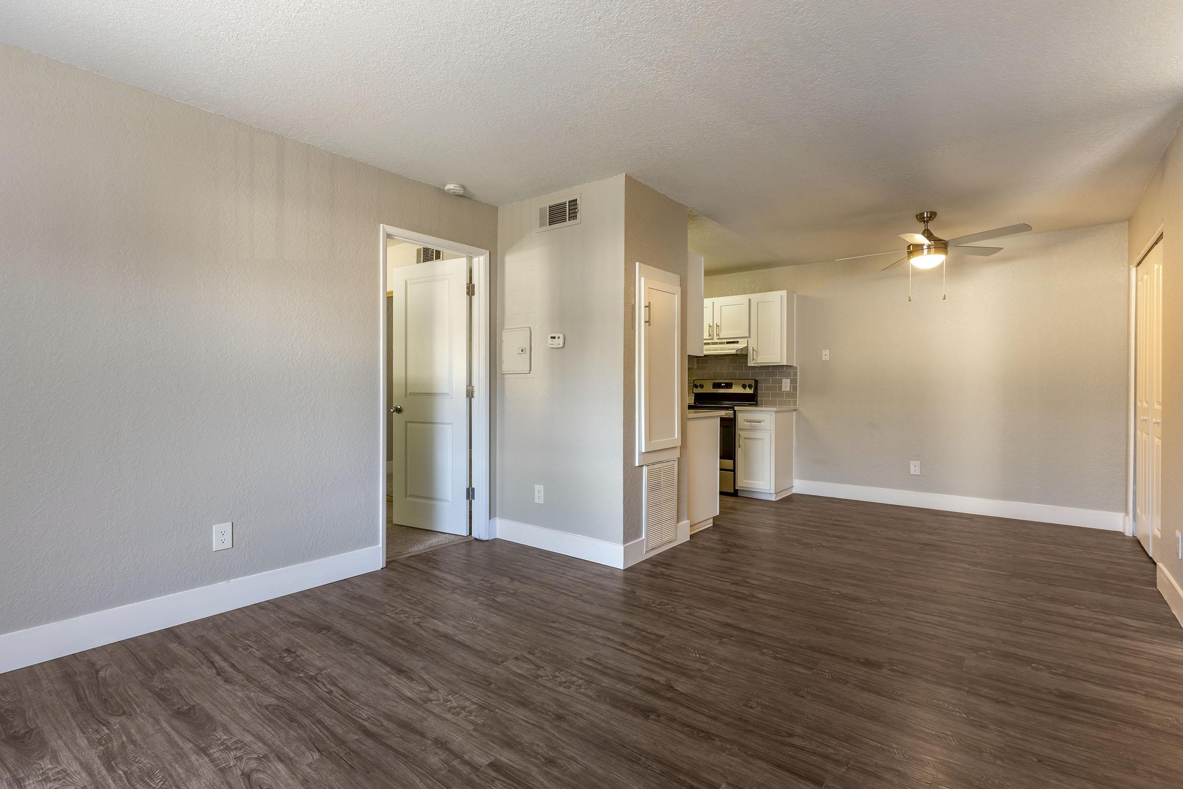 A spacious, light-colored living area with laminate flooring, featuring a ceiling fan, a doorway leading to another room, and an open kitchen in the background. Walls are painted in neutral tones, creating a bright and airy atmosphere.