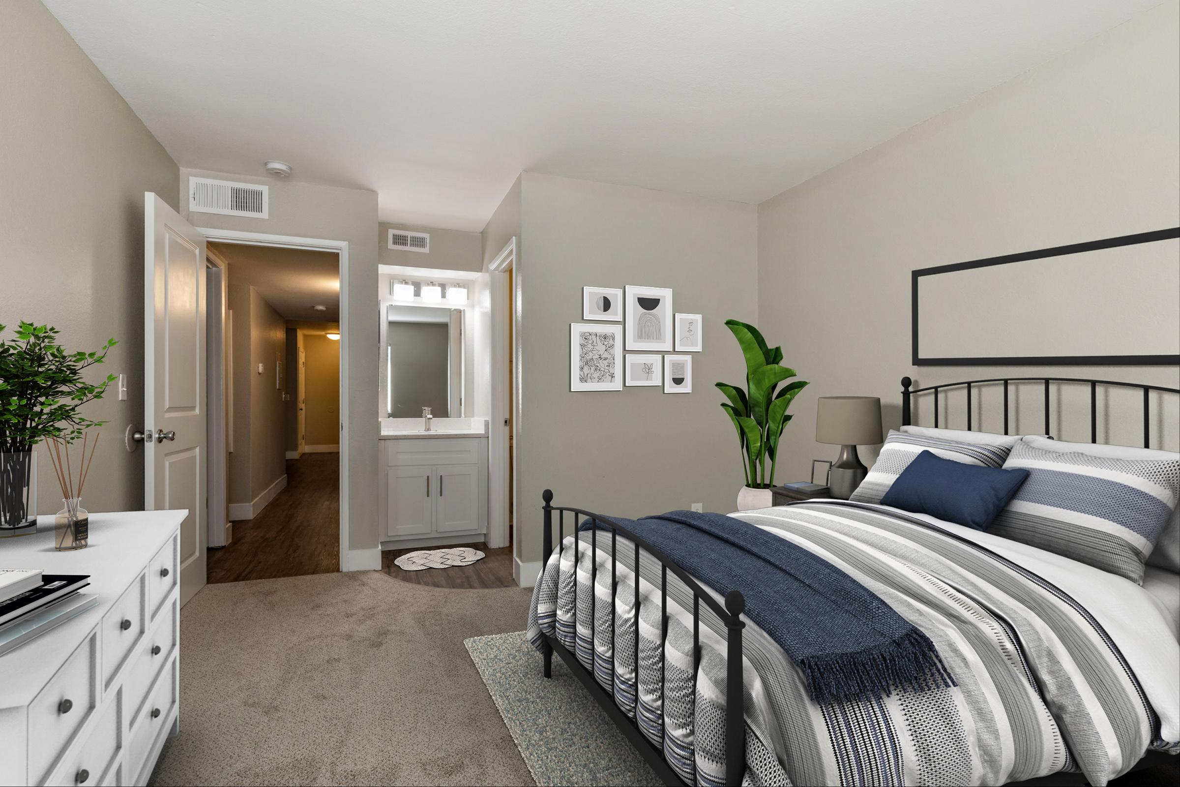 A cozy bedroom featuring a metal bed frame with gray and white bedding, a nightstand with a lamp, and a decorative plant. A small dresser is beside the bed. The room has an adjoining bathroom visible through an open door, with neutral walls and soft lighting. Framed artwork hangs on the walls.