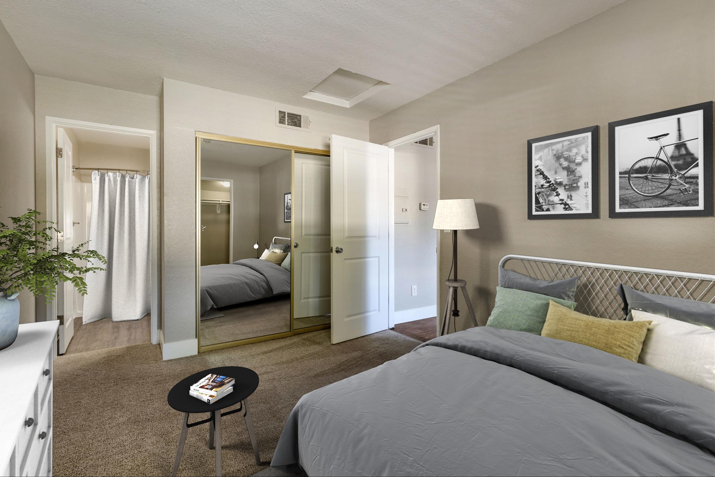 A cozy bedroom featuring light gray walls, a bed with gray linens and decorative pillows, a small black side table with a book, a white dresser, and a lamp. A mirrored closet reflects a glimpse of the room, while framed black and white photos adorn the wall. A curtain covers a nearby doorway.