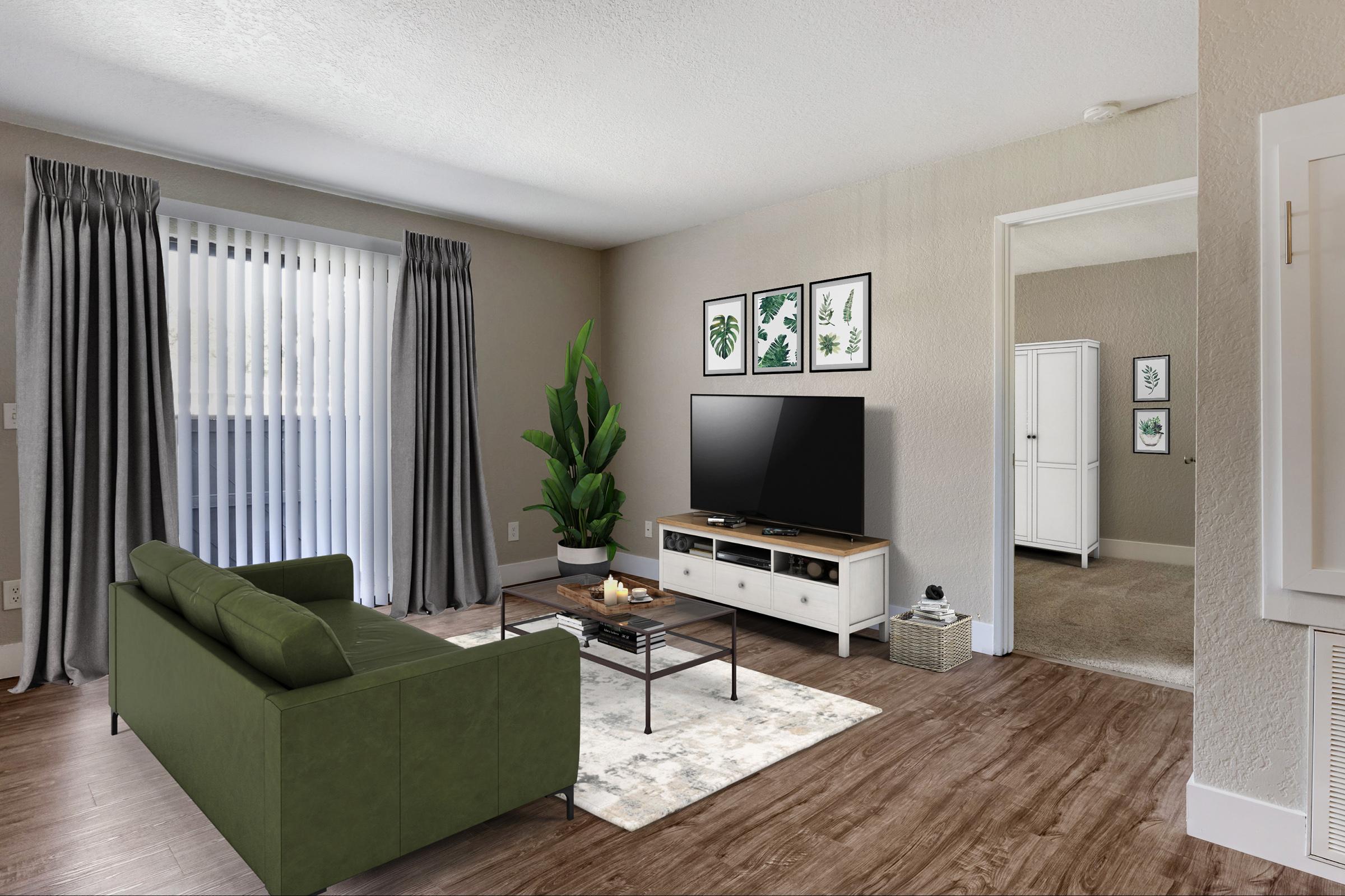 A modern living room featuring a green sofa, a coffee table with decor, and a TV on a white media console. The walls are light gray, adorned with framed botanical art. Large windows with sheer and solid curtains let in natural light, and a potted plant adds a touch of greenery to the space.