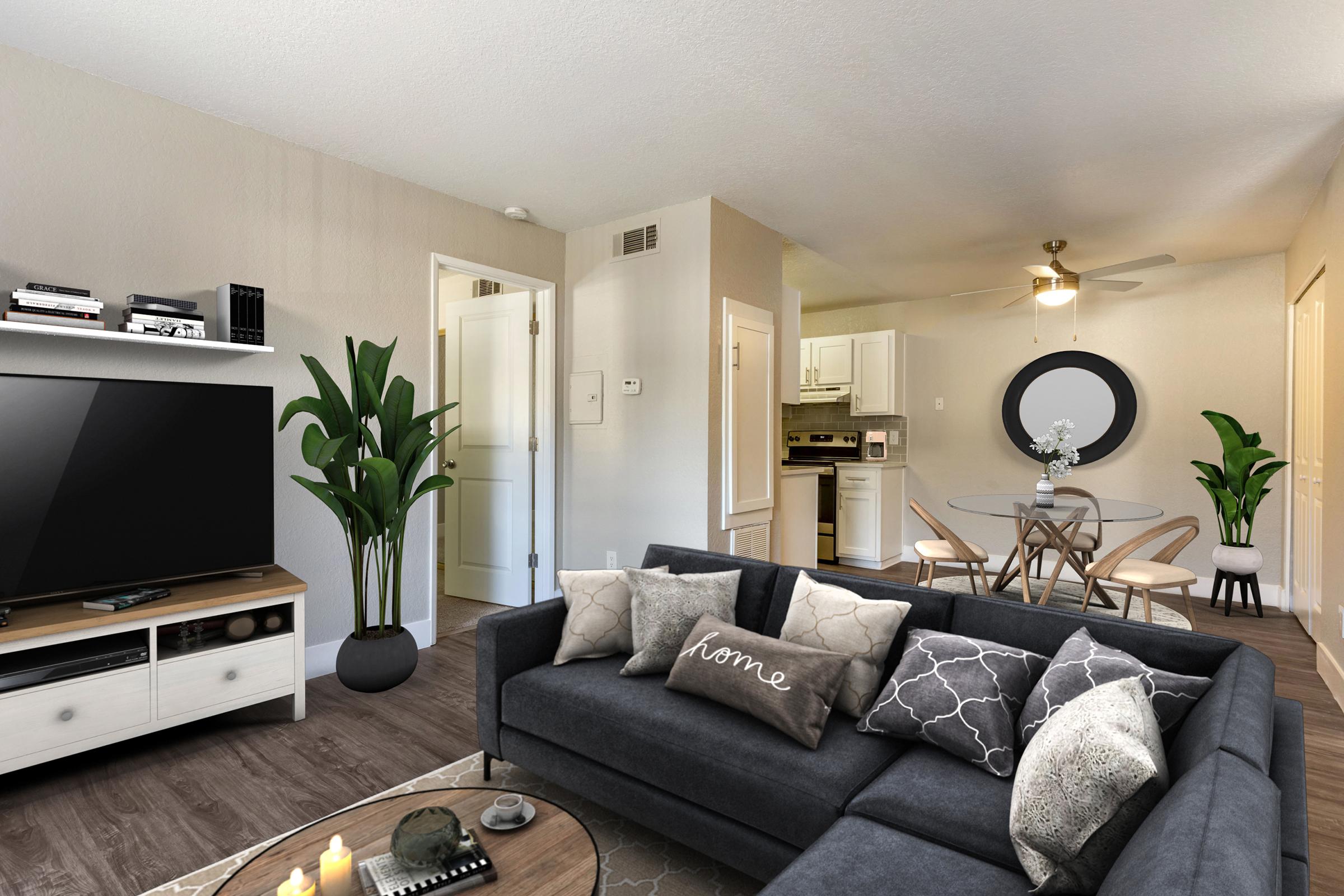 A modern, stylish living room featuring a dark gray sofa with decorative pillows, a coffee table with candles, a large flat-screen TV on a white entertainment unit, and potted plants. The space includes a dining area with a round table and chairs, and a light-colored kitchen visible in the background.