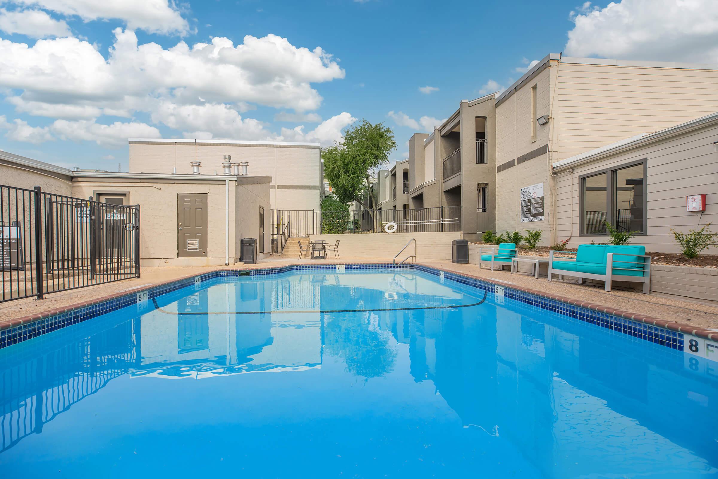 A clear blue swimming pool surrounded by a safety fence, with lounge chairs and tables nearby. The background features residential buildings under a partly cloudy sky.
