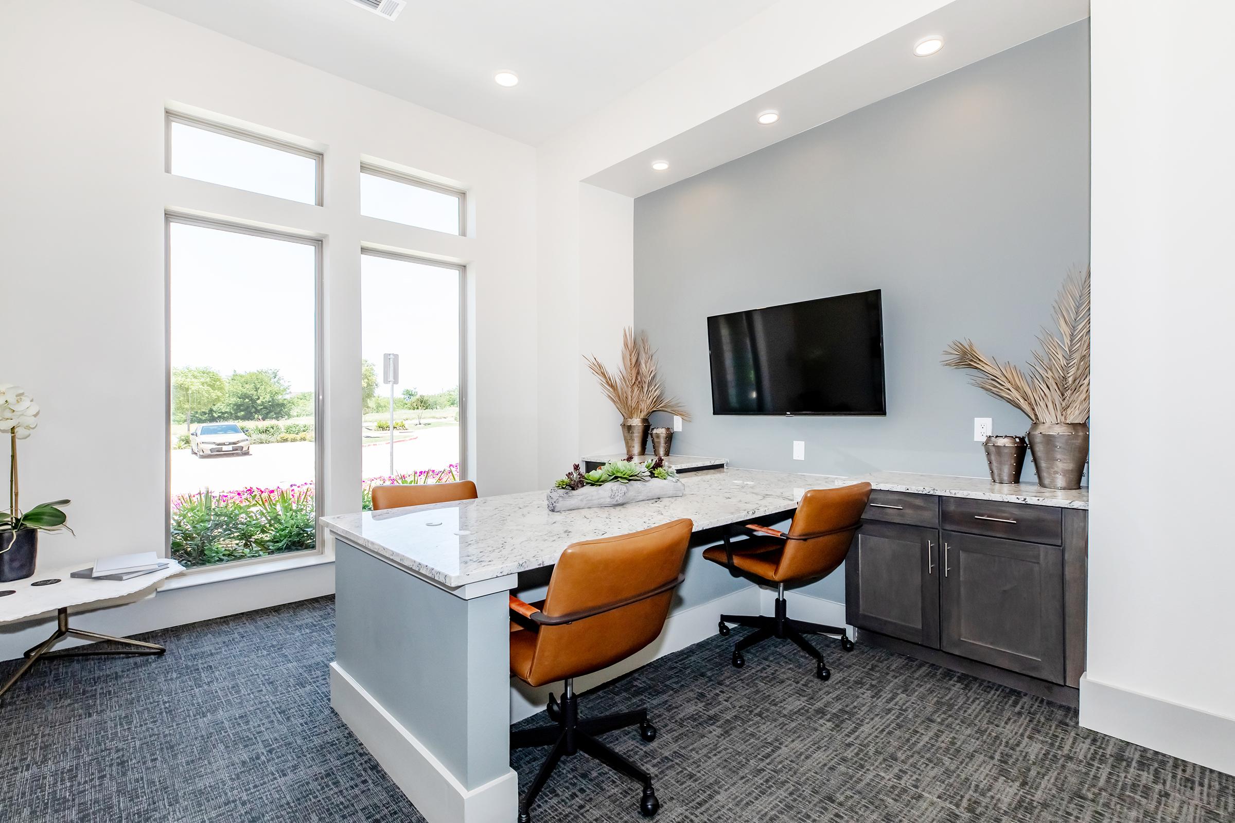 A modern office space featuring a workstation with a granite countertop, two brown chairs, and a wall-mounted TV. The room has large windows allowing natural light, with decorative plants and a stylish, minimalist design.