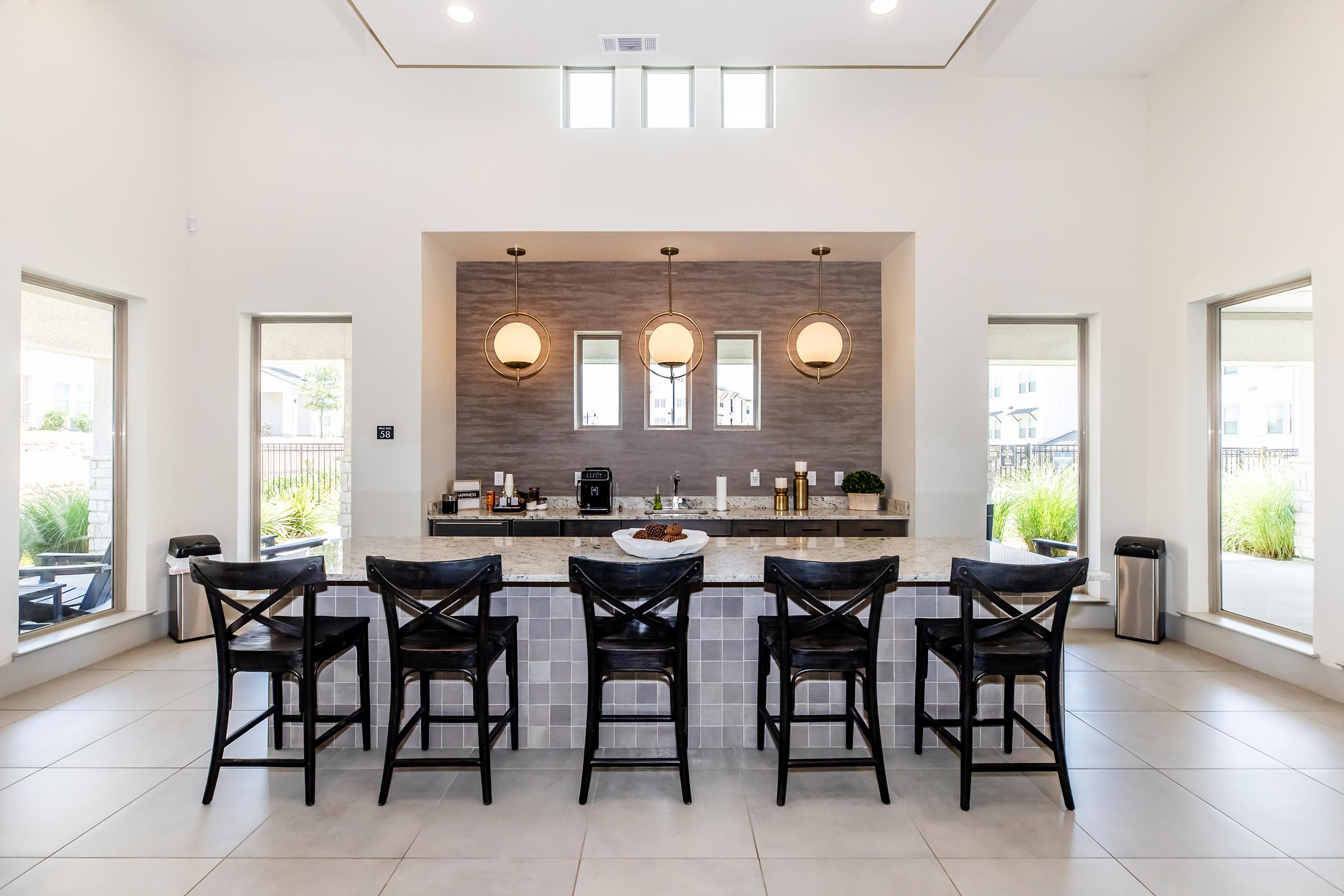 Modern kitchen interior featuring a large island with bar seating for six, surrounded by stylish black chairs. The space is well-lit with large windows on either side, and decorative pendant lights hanging above the island. Clean lines and neutral colors create a contemporary atmosphere.
