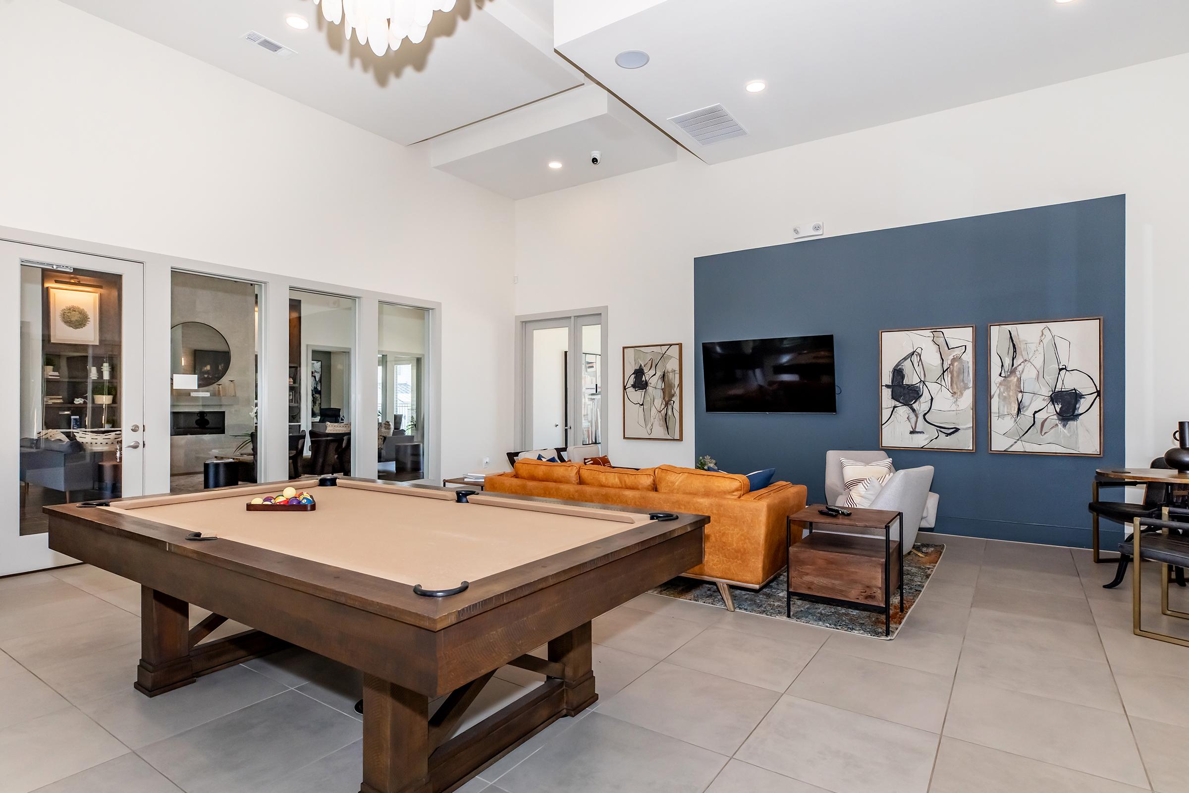 Modern interior living space featuring a billiards table in the center, a cozy orange sofa, and a dark blue accent wall with framed artwork. Large glass doors lead to an outdoor area, and a flat-screen TV is mounted on the wall. The room has neutral tile flooring and stylish decor, creating a comfortable atmosphere.