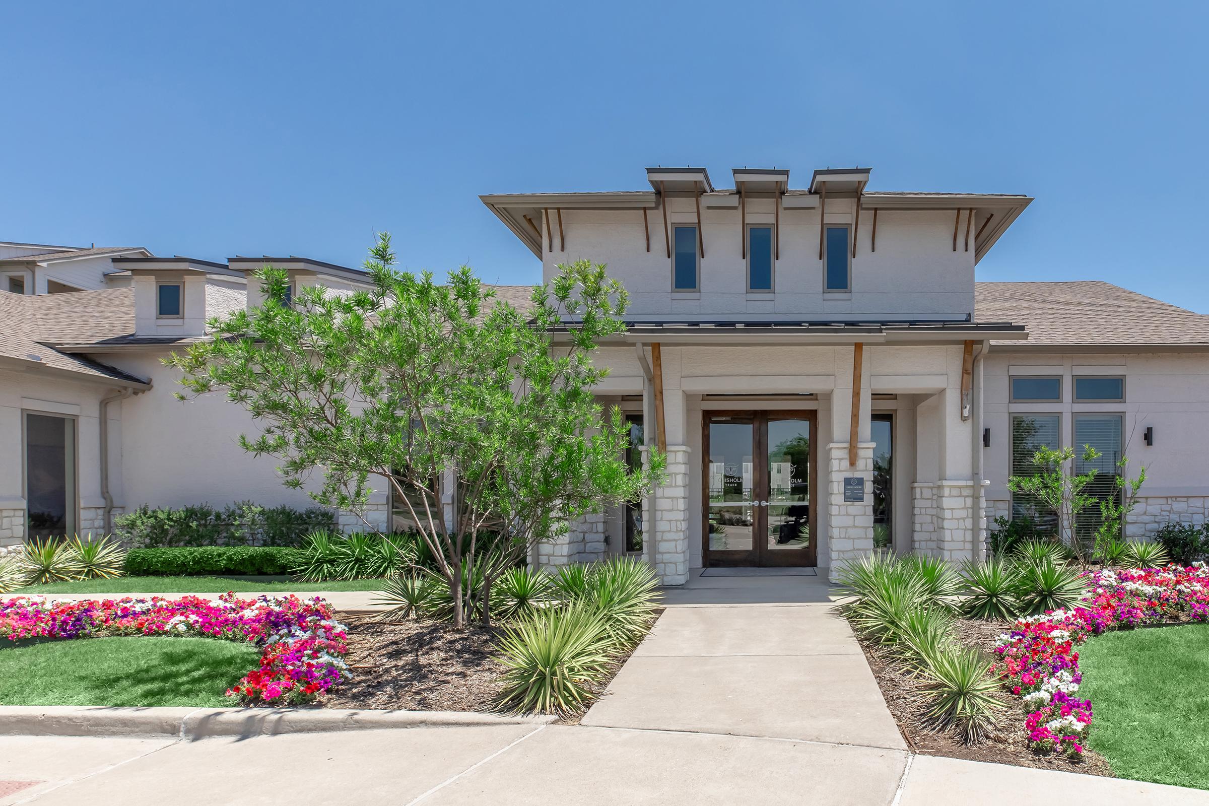 A modern residential building with a welcoming entrance. The facade features cream-colored walls and a wooden accent above the door. Vibrant flower beds with pink and purple flowers surround the entrance, complemented by neatly landscaped greenery under a clear blue sky.