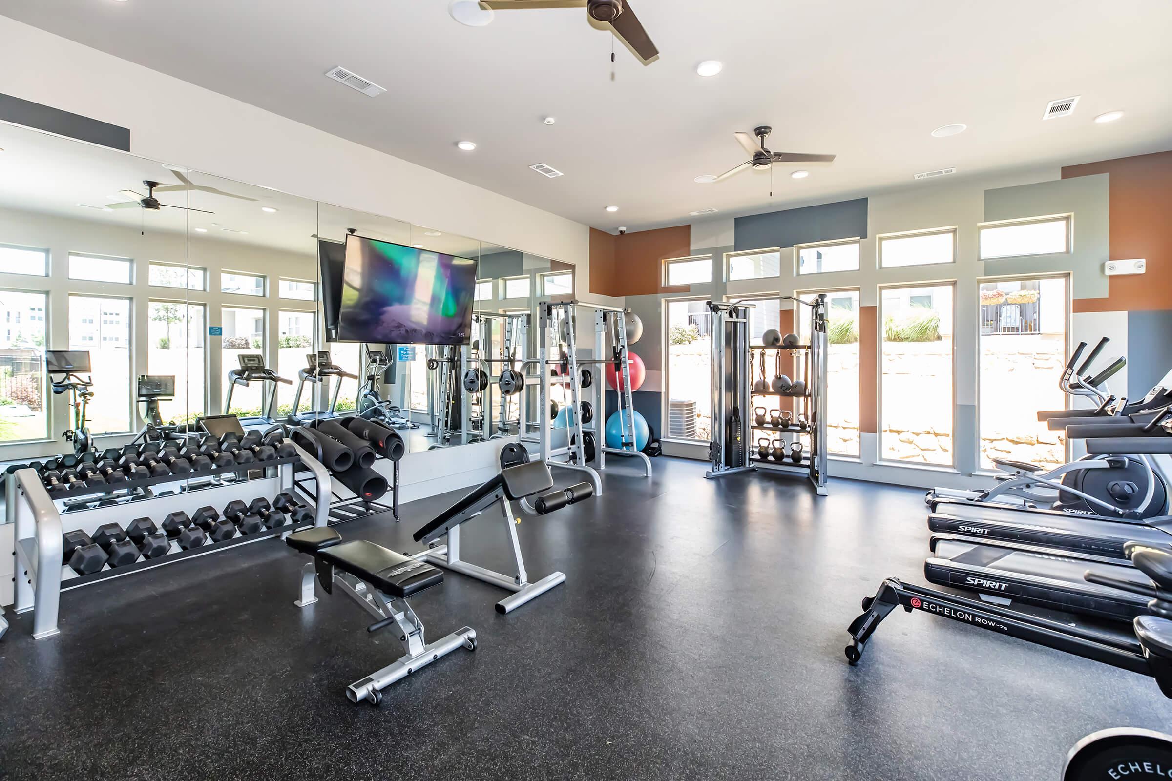 A bright and spacious gym with large windows, featuring various workout equipment including free weights, benches, treadmills, and a flat-screen TV. The room has a modern design with colorful accent walls and is well-lit, creating an inviting atmosphere for fitness activities.