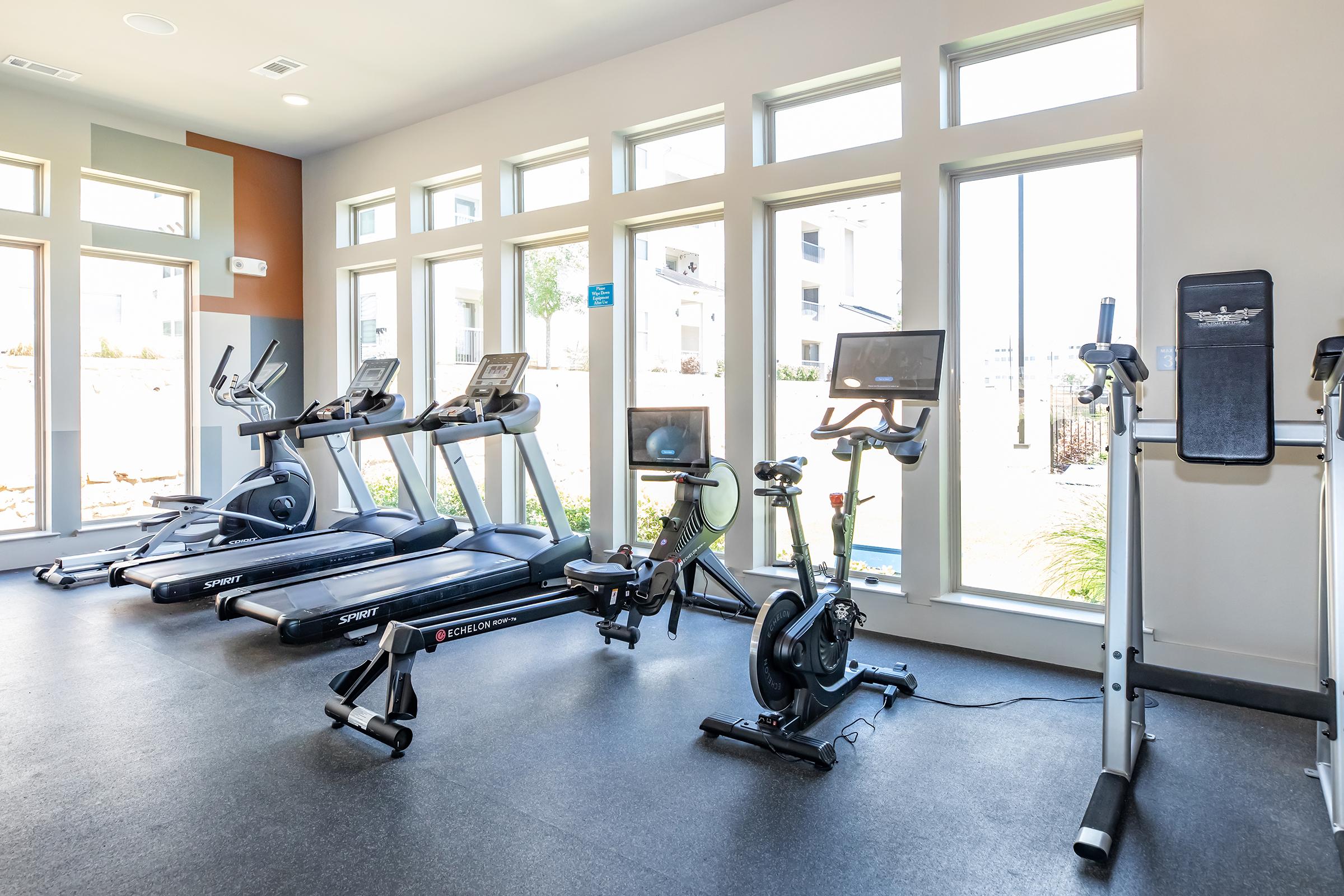 A modern fitness room featuring multiple cardio machines including treadmills and a stationary bike. Large windows allow natural light to fill the space, creating a bright and inviting atmosphere. The room is well-maintained with a sleek, contemporary design.