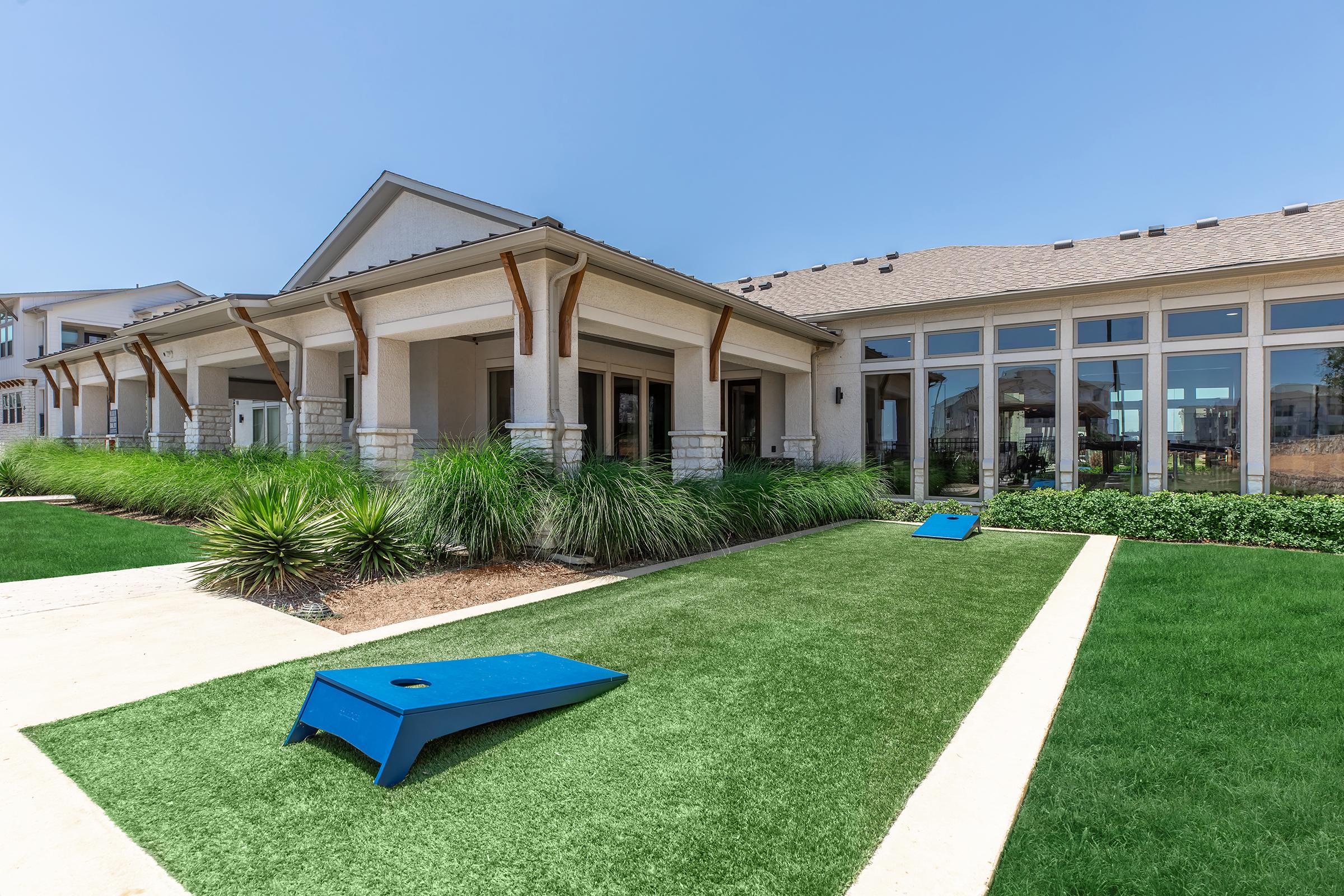 A well-maintained outdoor area featuring a green lawn with two blue cornhole boards set up for play. In the background, a stylish building with large windows and a porch is visible, surrounded by landscaped grass and plants, under a clear blue sky.