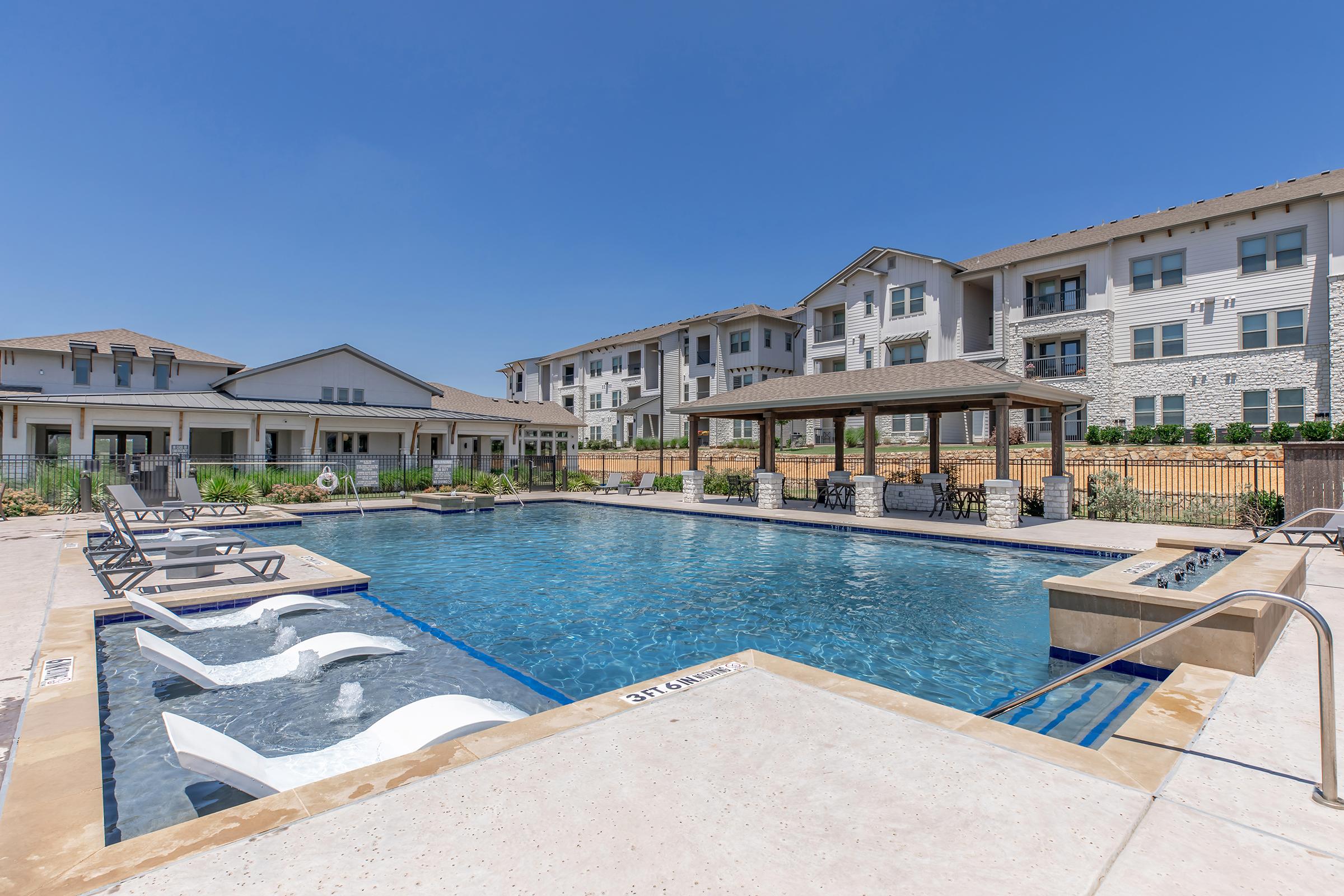 A clear blue swimming pool surrounded by lounge chairs, with a shaded gazebo area in the background. Modern apartment buildings are visible on the right side, and a well-maintained landscape complements the recreational facilities under a clear blue sky.