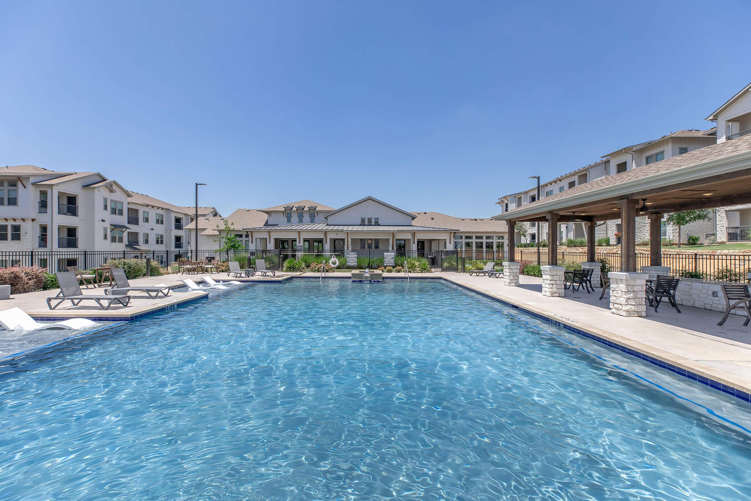 A sparkling blue swimming pool surrounded by lounge chairs and shaded areas, with modern residential buildings in the background. The sky is clear and sunny, creating a relaxed outdoor atmosphere ideal for leisure and relaxation.