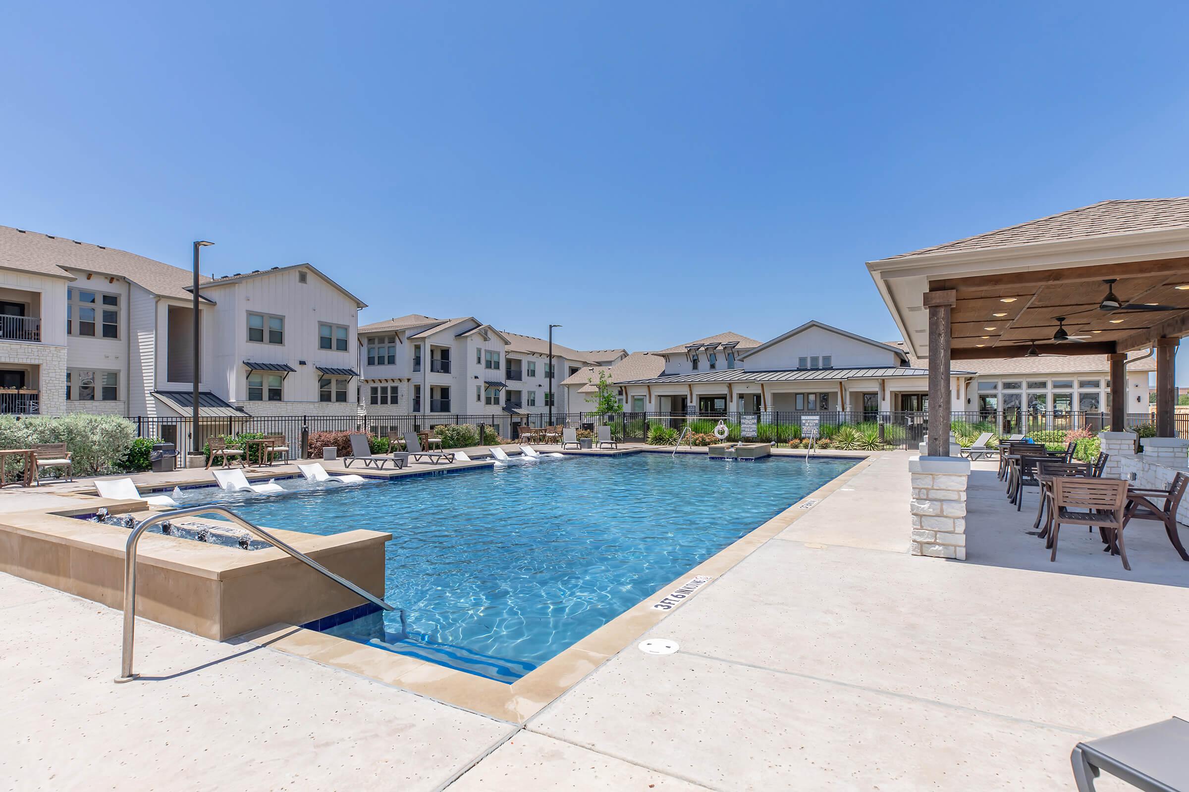 Swimming pool area with modern lounge chairs and a shaded seating area nearby. Surrounding the pool are well-maintained landscaping and multi-story residential buildings in a sunny environment, creating a relaxed atmosphere for outdoor leisure.