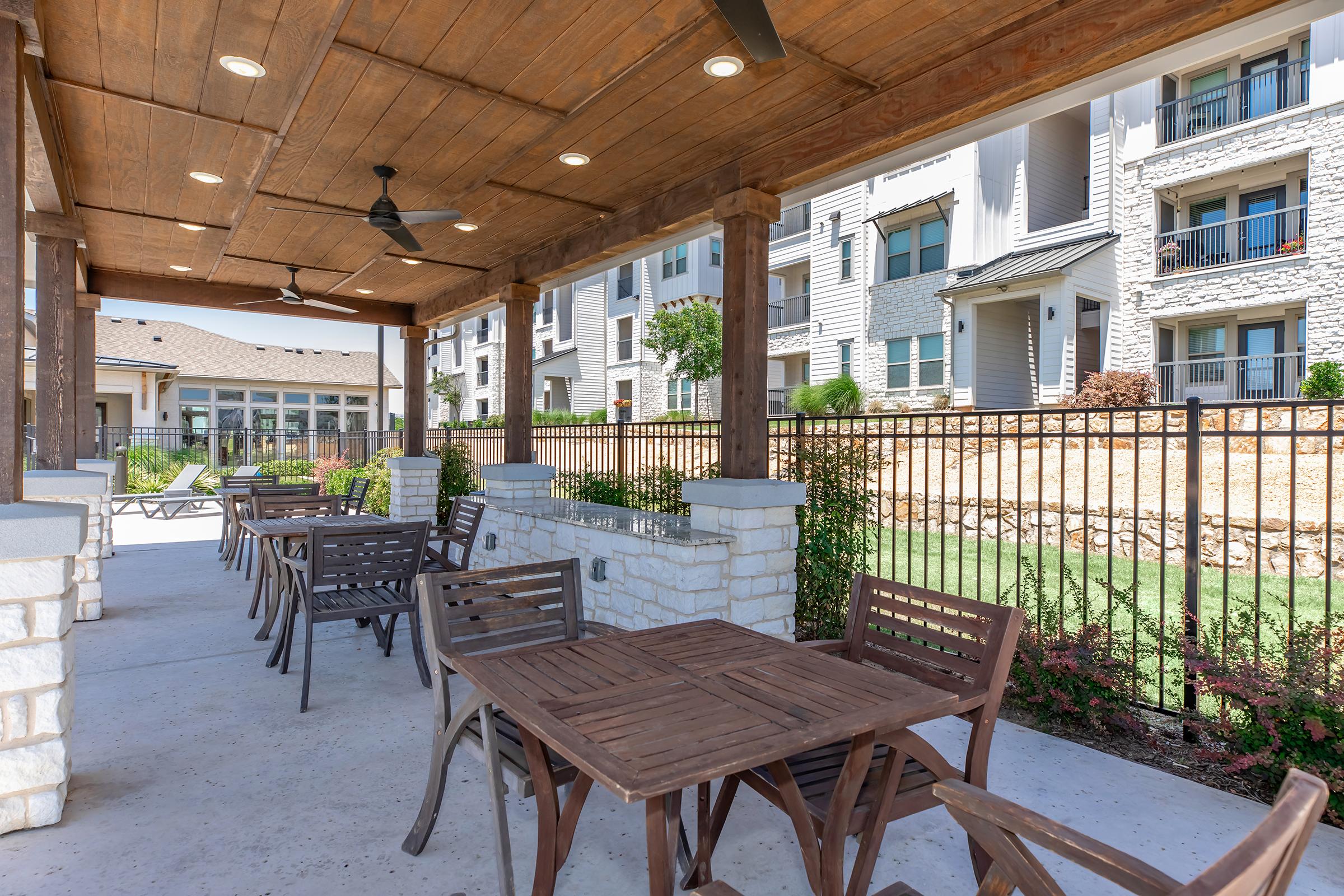 Outdoor seating area with wooden tables and chairs under a covered patio. The space features ceiling fans and is surrounded by a stone wall and decorative fencing. In the background, modern apartment buildings are visible, along with landscaped greenery. Bright, sunny day creating a welcoming atmosphere.