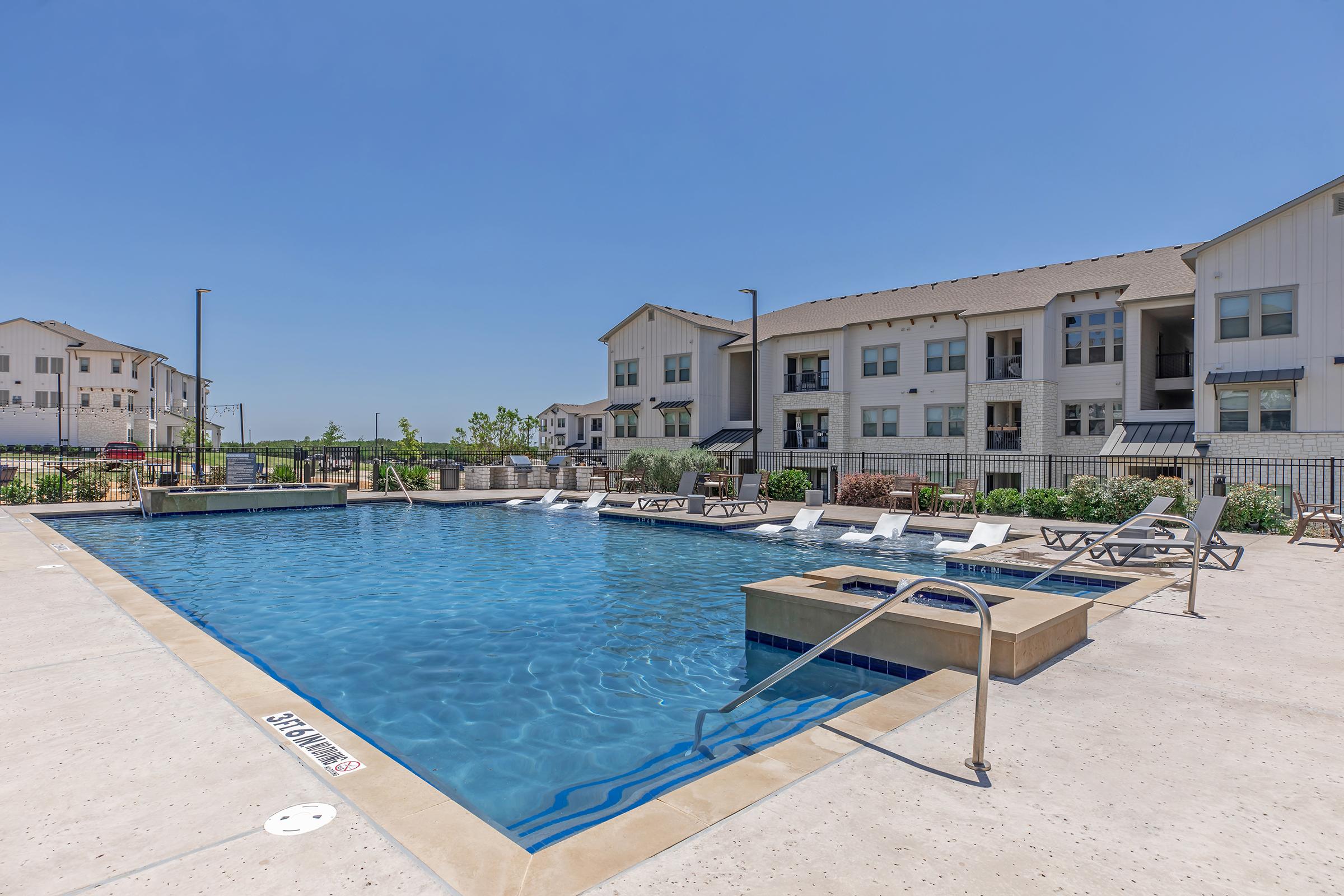 A modern outdoor swimming pool surrounded by lounge chairs and landscaped greenery. In the background, several multi-story residential buildings are visible under a clear blue sky. The pool area is well-maintained, offering a relaxing environment.