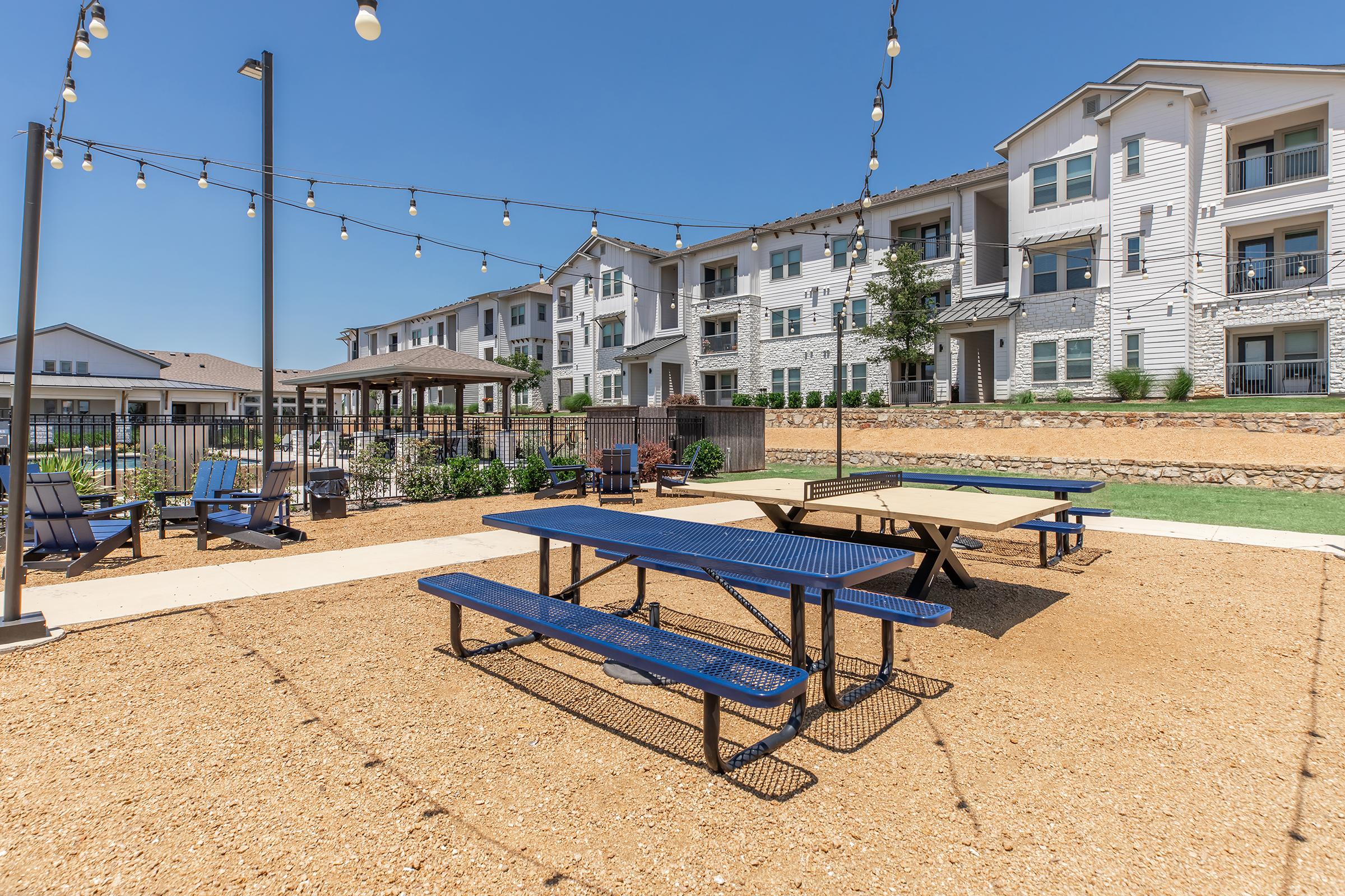 Outdoor recreational area featuring picnic tables and a ping pong table on a gravel surface, surrounded by green landscaping and modern apartment buildings. String lights hang above, creating a welcoming atmosphere for gatherings and leisure activities.