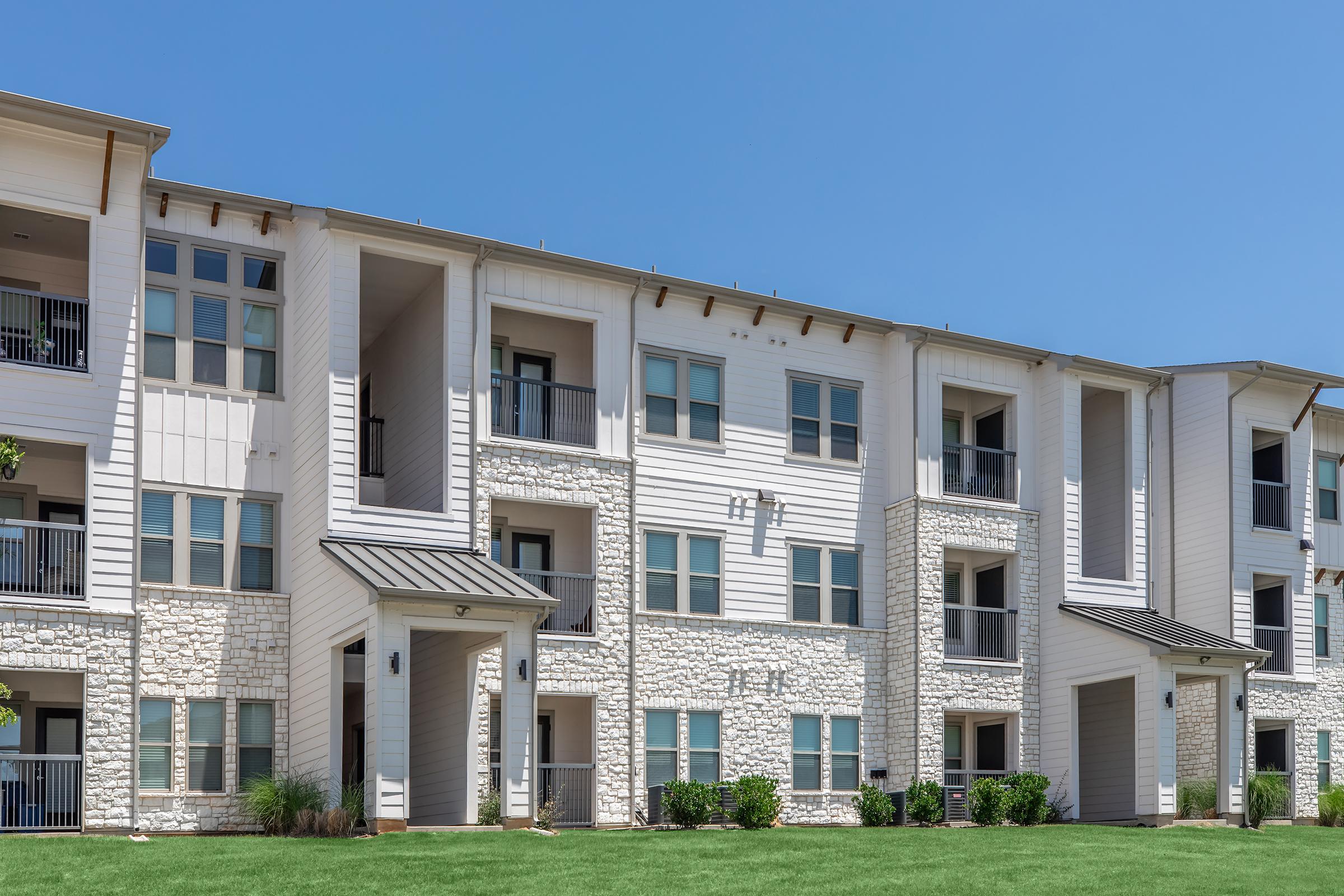 Modern apartment building with multiple balconies and stone accents, surrounded by green grass under a clear blue sky. The structure features a light-colored exterior and large windows, showcasing a contemporary architectural design.