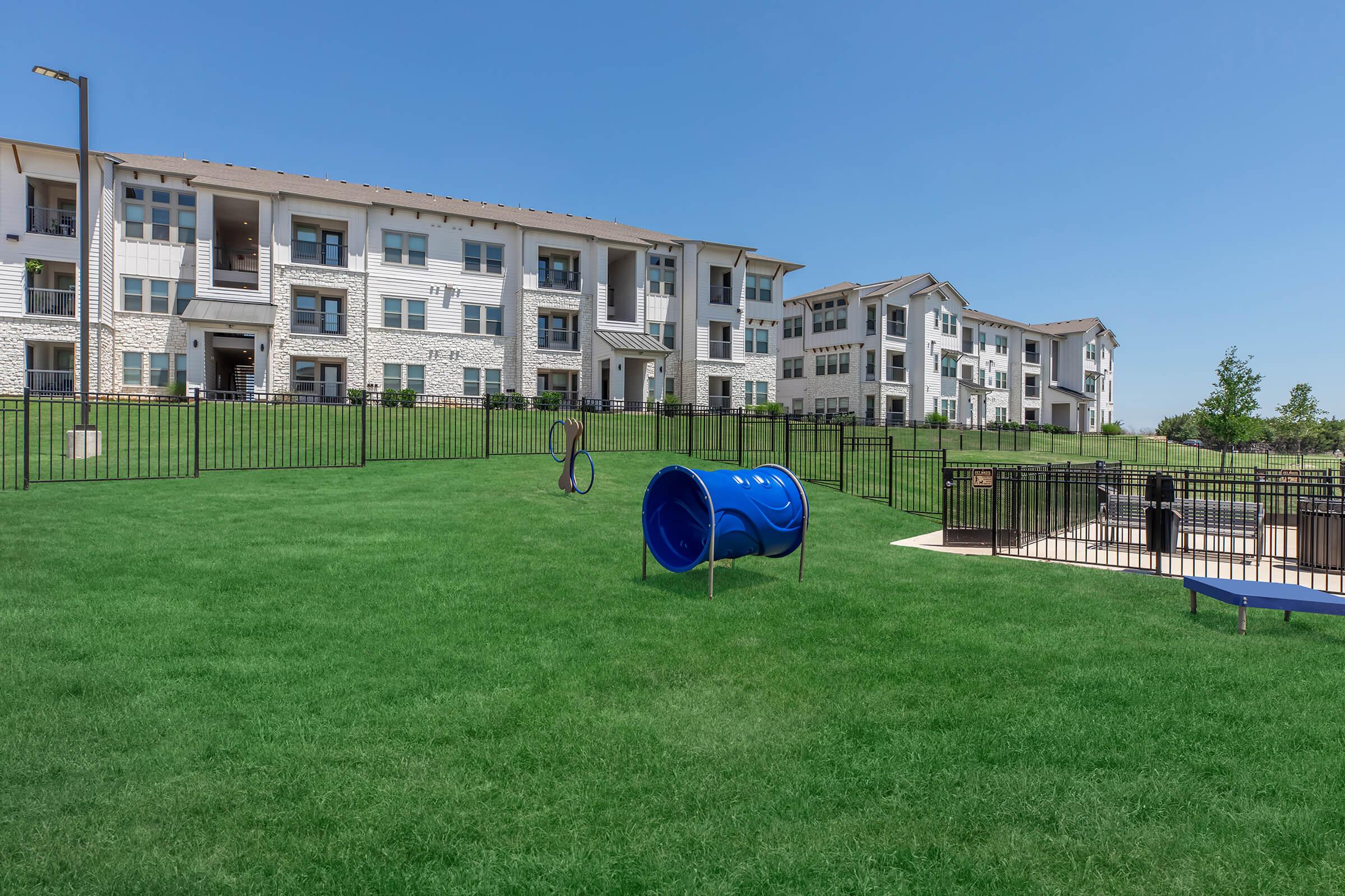 A well-maintained grassy area in front of modern apartment buildings, featuring a blue playground tube and a fenced pool area. The sky is clear and sunny, highlighting the greenery and the spacious layout of the outdoor space.