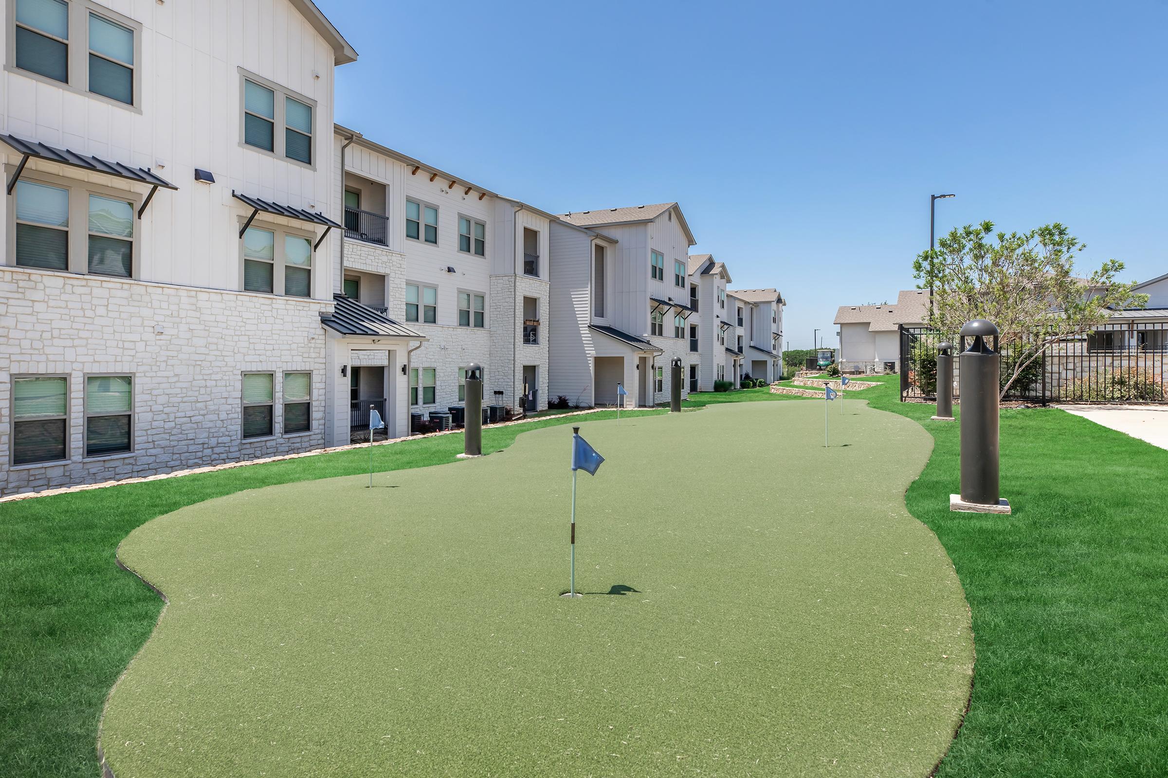 A landscaped putting green with several golf flags is situated in a residential complex. Surrounding the green, there are modern apartment buildings with a mix of white stone and siding. The scene is set under a clear blue sky, with well-maintained greenery enhancing the outdoor space.