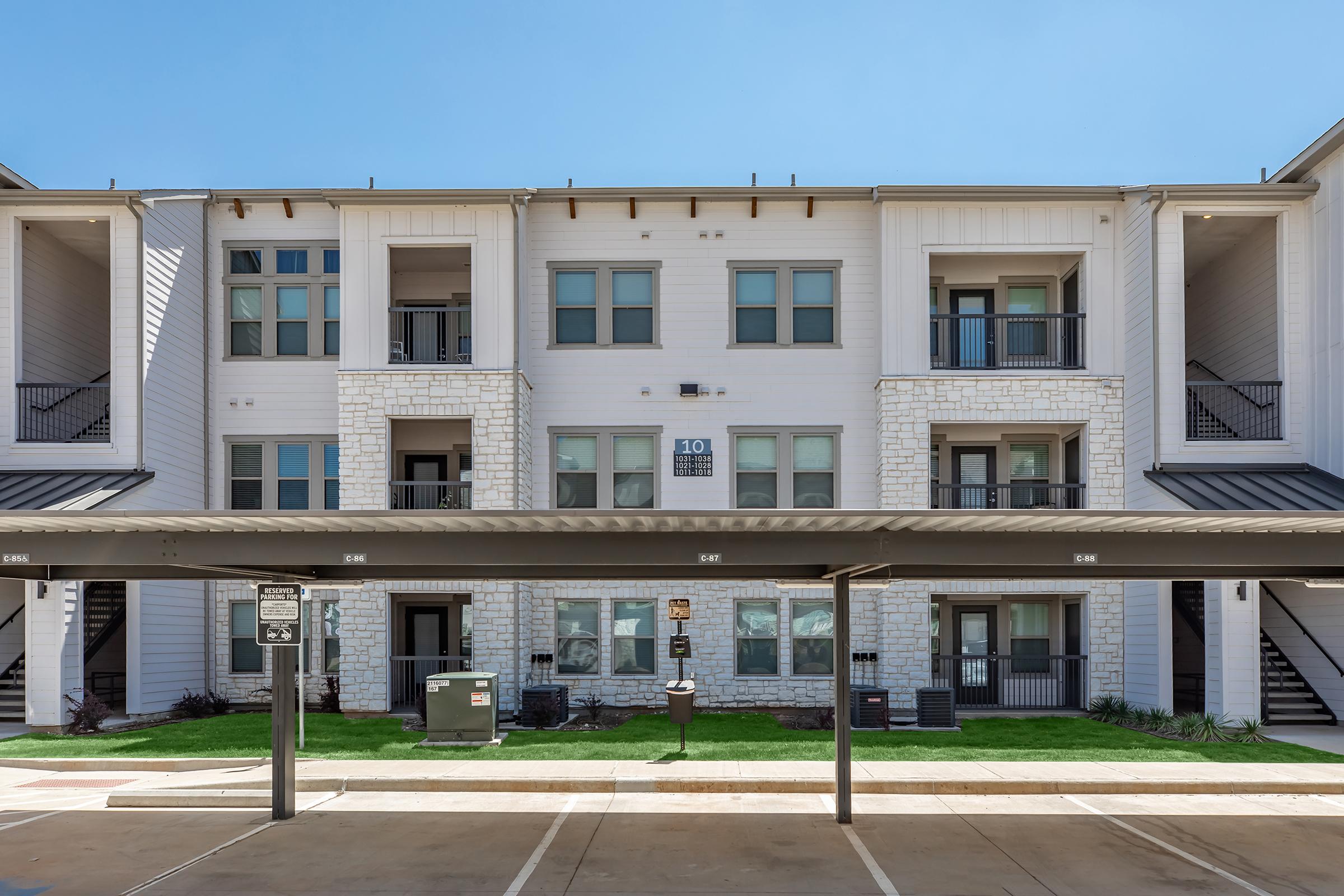 Three-story residential apartment building featuring a modern design, with a white exterior and stone accents. Balconies can be seen on each level, alongside covered parking spots in the foreground. The scene is bright with clear blue skies and manicured landscaping.