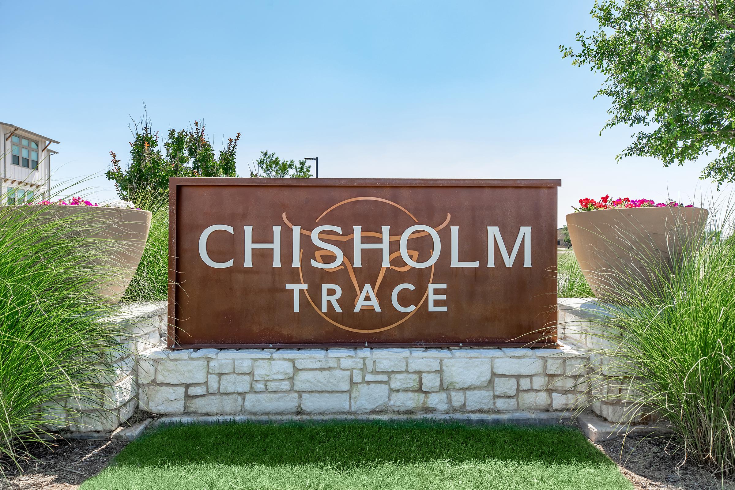 Sign for Chisholm Trace with a modern design, featuring large white lettering on a rust-colored metal background, flanked by decorative planters with pink flowers. The sign is set against a clear blue sky and green landscaping, creating a welcoming entrance.