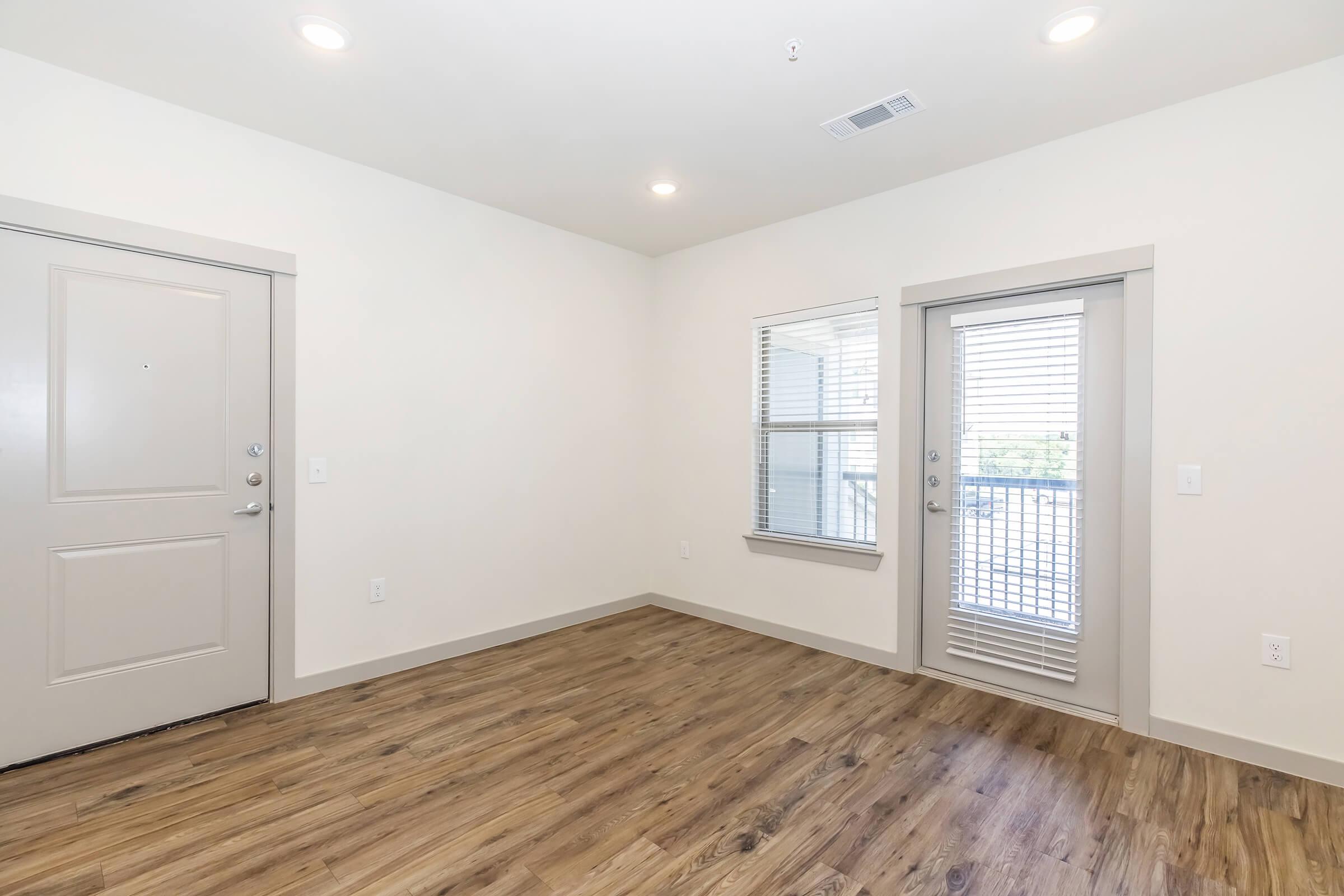 A modern, unfurnished room featuring light-colored walls and wooden flooring. There is a single door leading outside, a large window with blinds, and ample natural light illuminating the space. The room is clean and spacious, making it suitable for various purposes such as a living area or office.