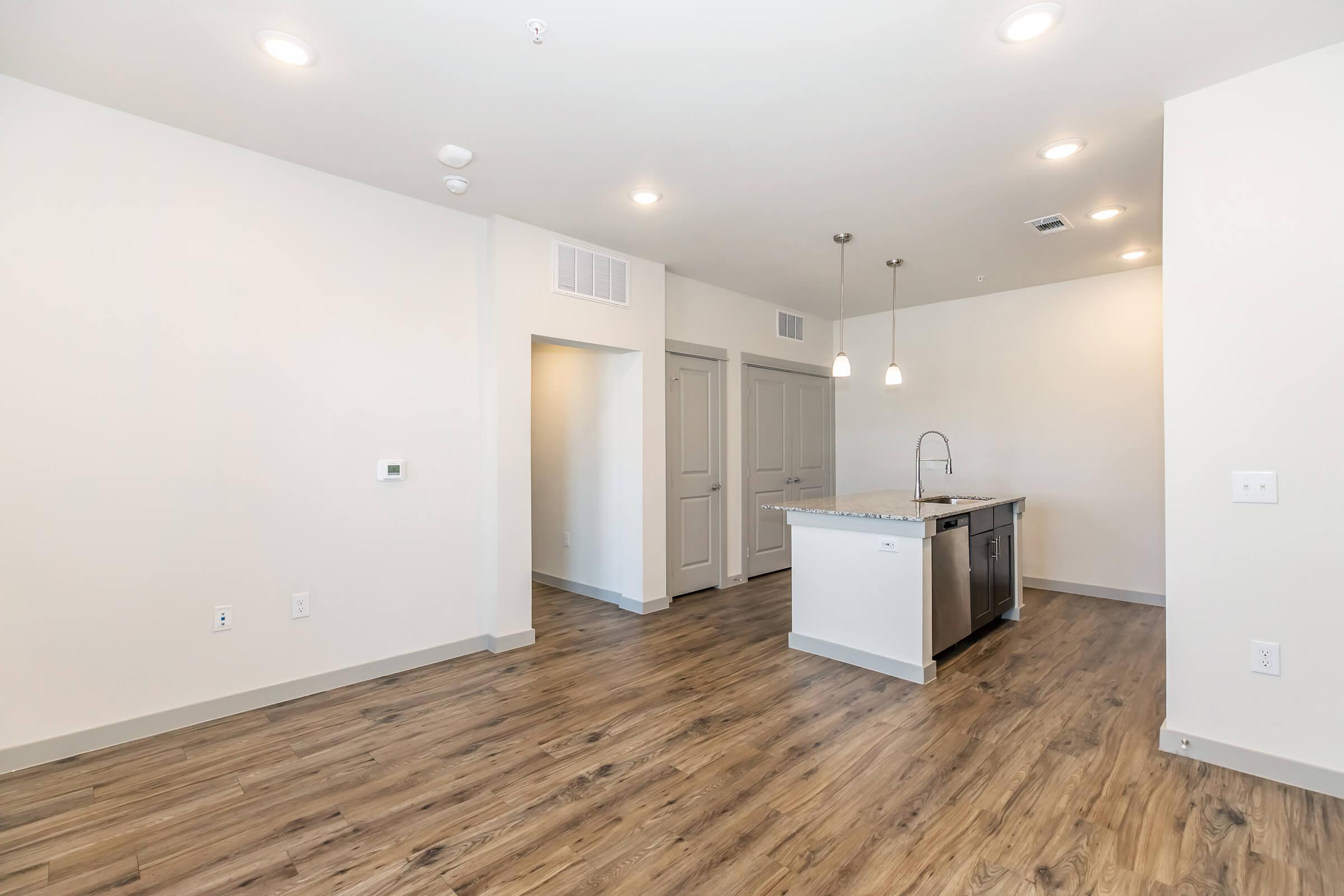 Interior view of a modern apartment with an open floor plan. The space features a kitchen with a central island and stainless steel appliances, light wood-like flooring, and neutral-colored walls. There are recessed ceiling lights providing ample illumination, and a doorway leading to another room.