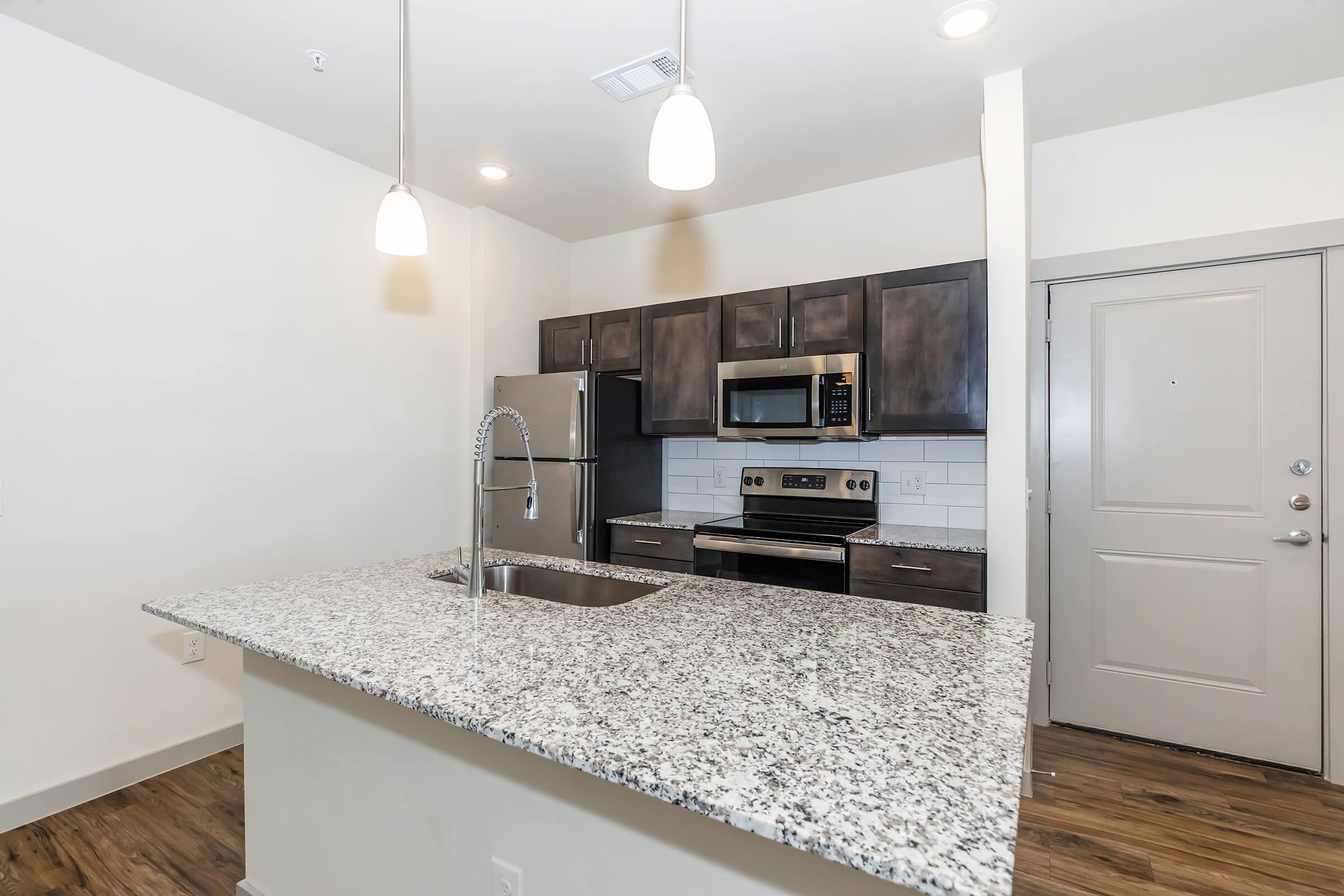 Modern kitchen featuring granite countertops, stainless steel appliances including a refrigerator and oven, dark cabinetry, and pendant lighting. The open layout connects to a living space, with a door visible on the right side leading outside. The flooring is wood-like laminate.