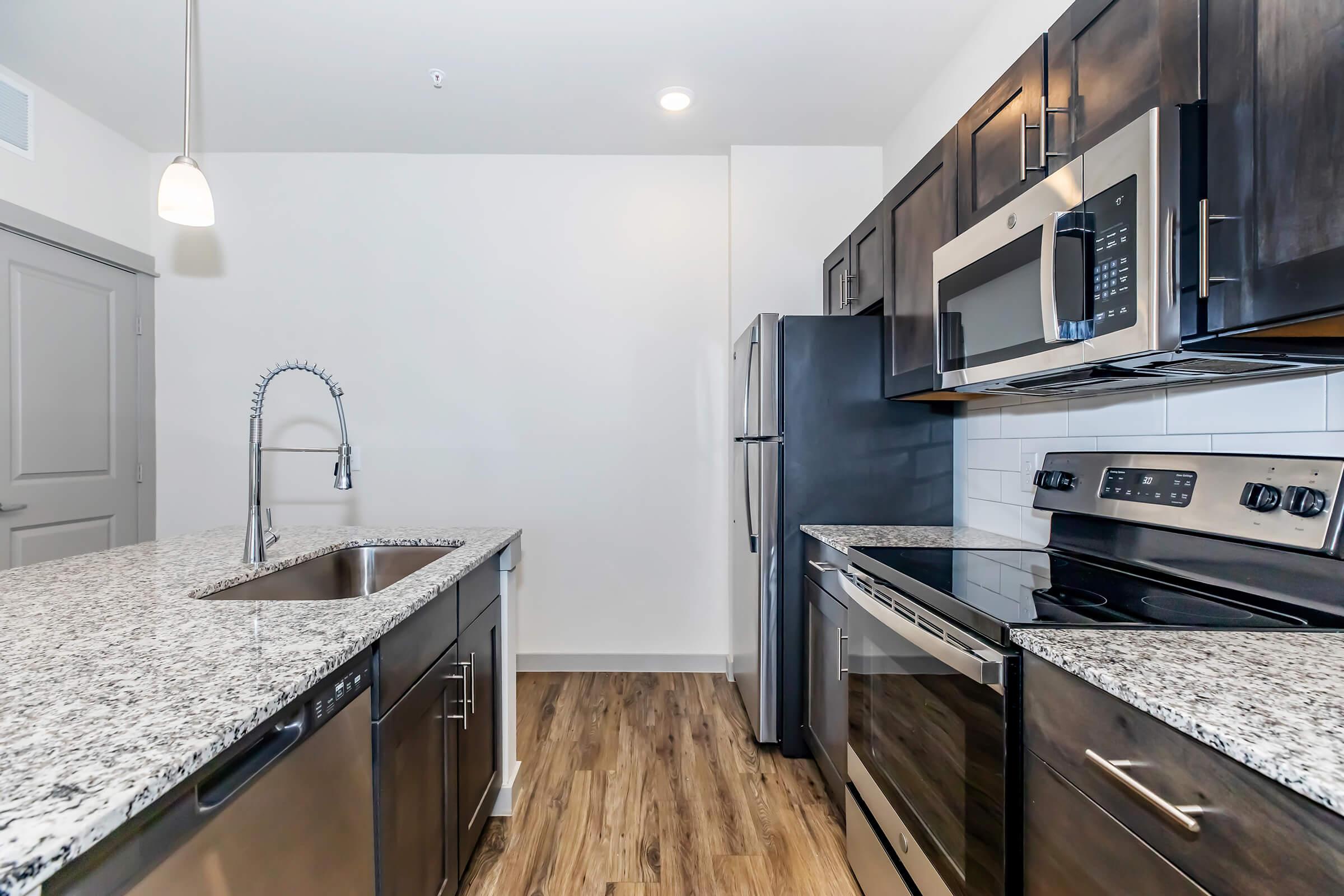 Modern kitchen featuring dark cabinetry, granite countertops, stainless steel appliances including a refrigerator, microwave, and oven. The kitchen island has a sink and a contemporary faucet, with a light fixture above. The flooring is wooden, and the walls are painted light colors, creating a bright and inviting space.