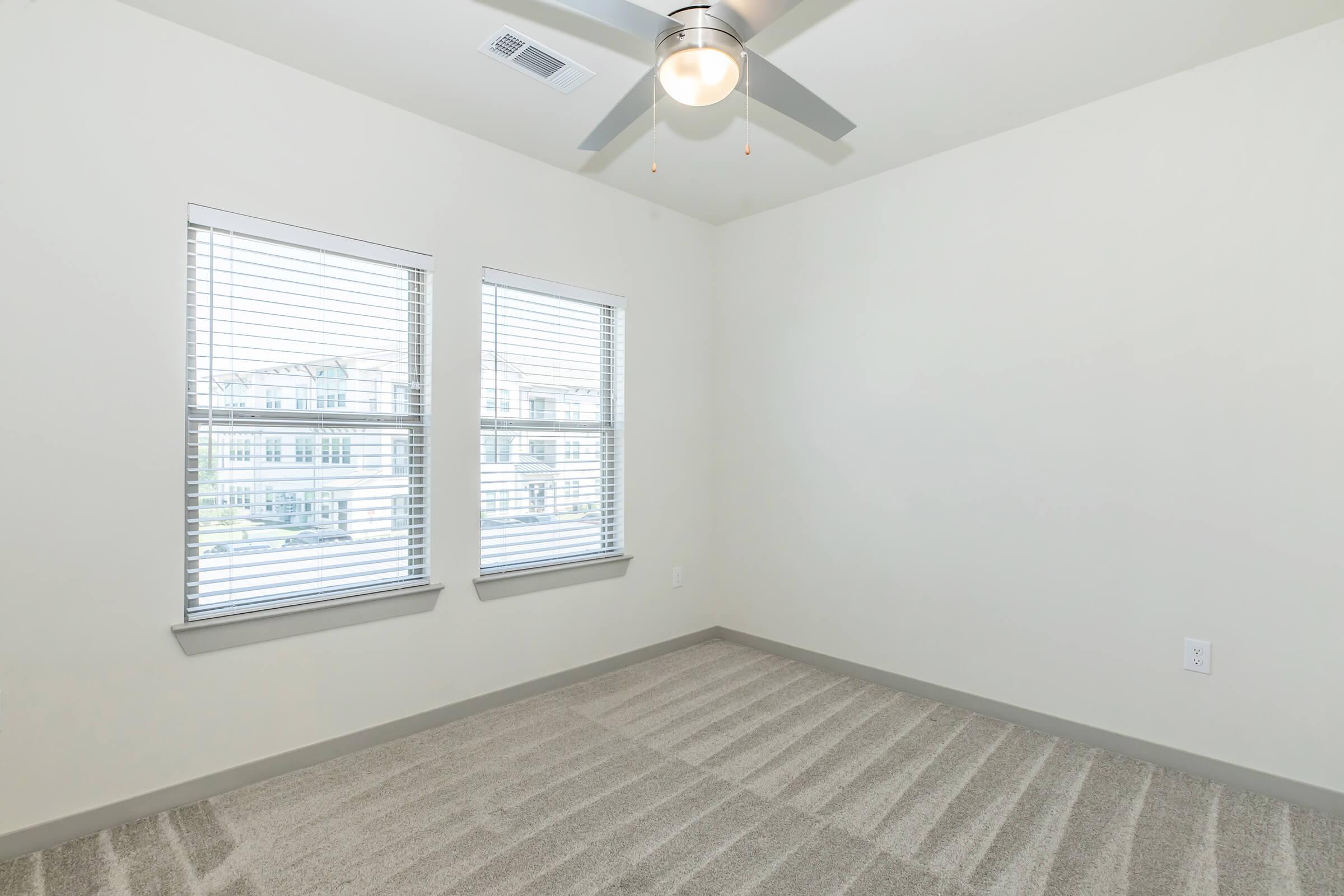 A vacant room with light-colored walls and carpet. It features two large windows with blinds, allowing natural light to enter. A ceiling fan is mounted above, and the room has a simple, modern design.